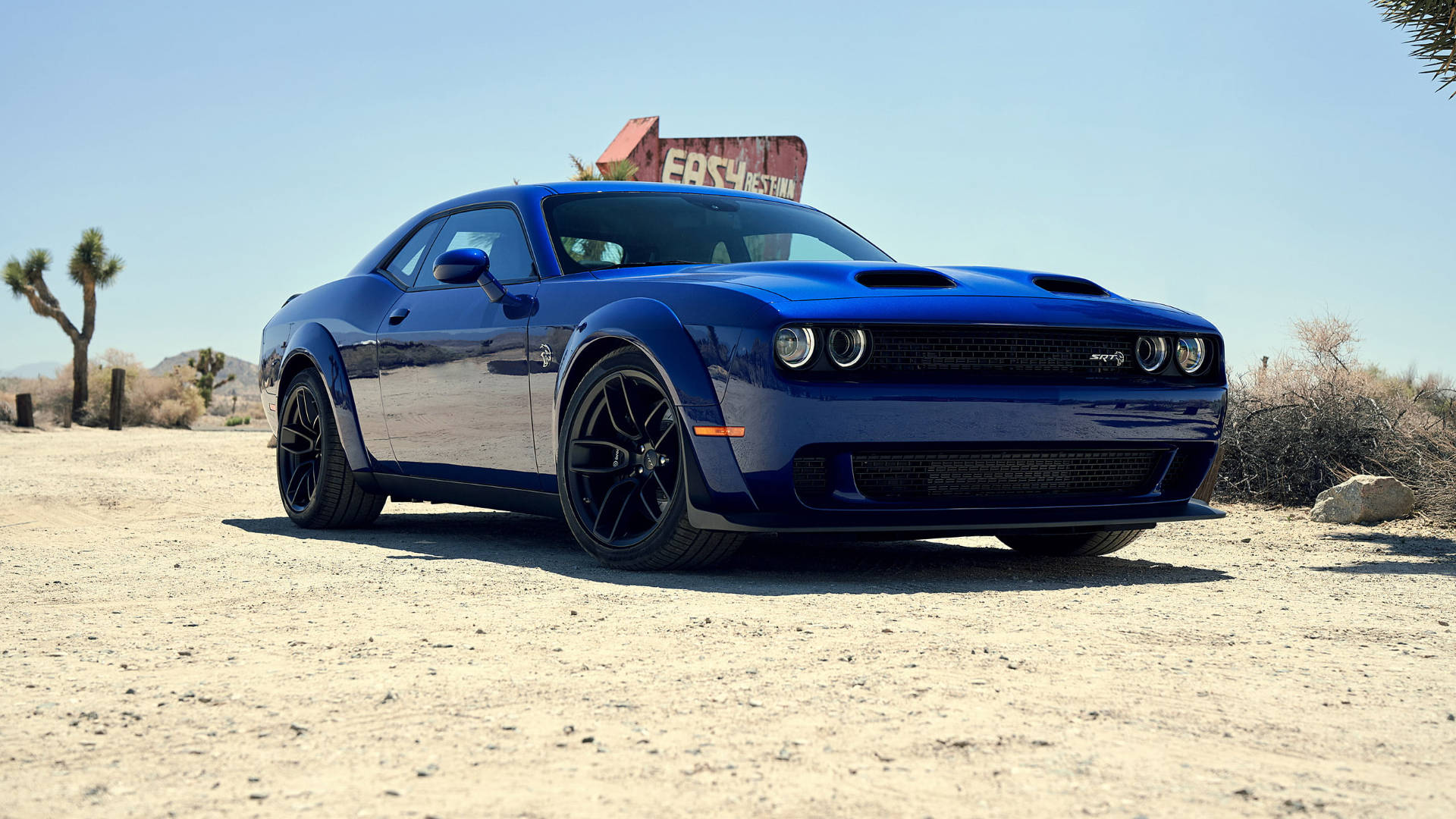 沙漠 桌布 壁紙 — 一架閃亮的藍色 Dodge Challenger 肌肉車停泊在塵土飛揚的沙漠道路上，背景是晴朗的藍天和一個褪色的路邊標誌。