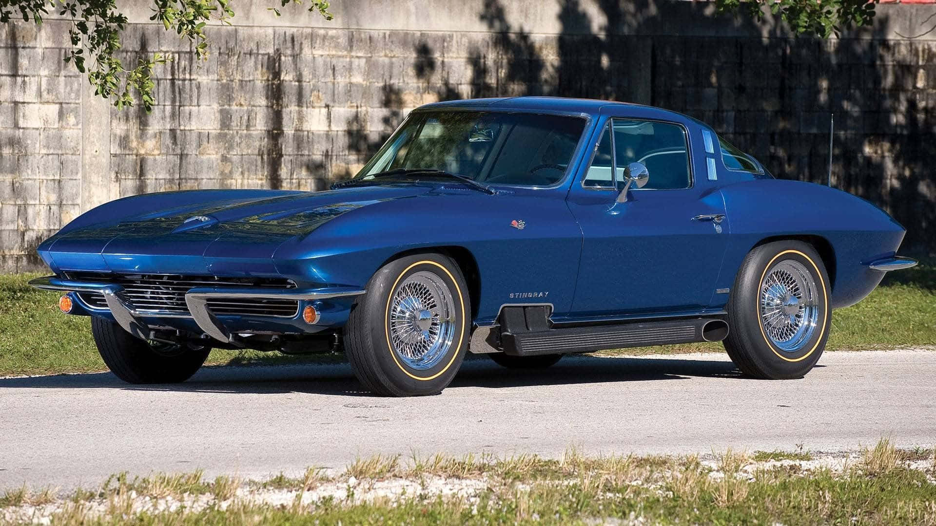 classic 桌布 壁紙 — 一架閃亮藍色經典Corvette Stingray，配備鍍鉻鋼絲輪圈，停泊在混凝土牆和草地旁的鋪砌道路上。