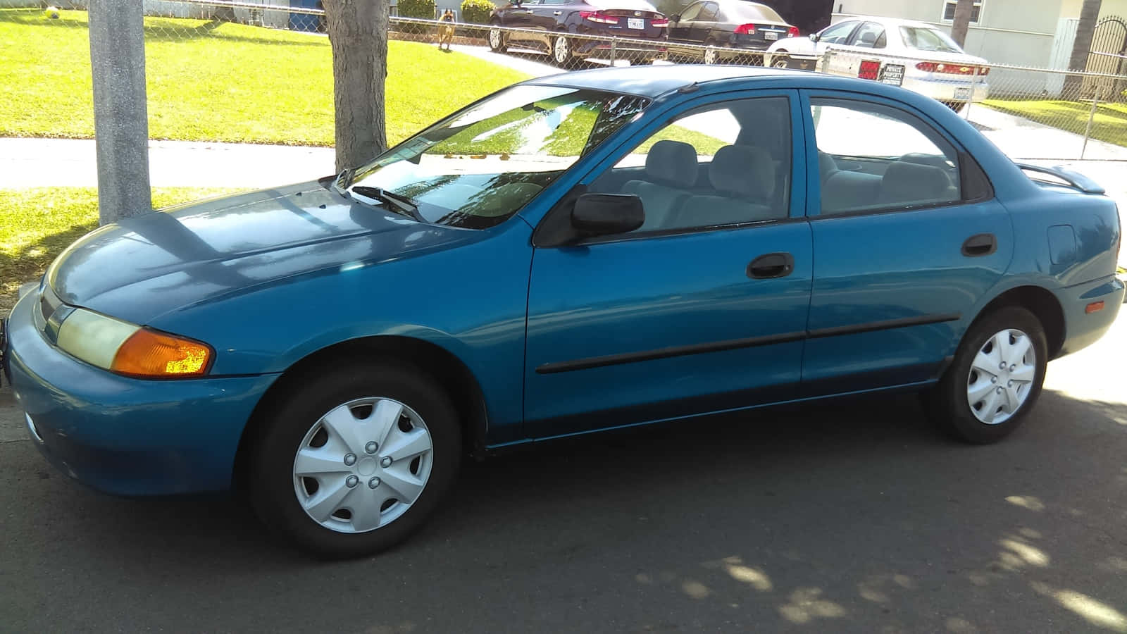 Mazda Protege 桌布 壁紙 — 一架青綠色房車停泊在陽光明媚的街道上，背景有樹木和其他車輛。