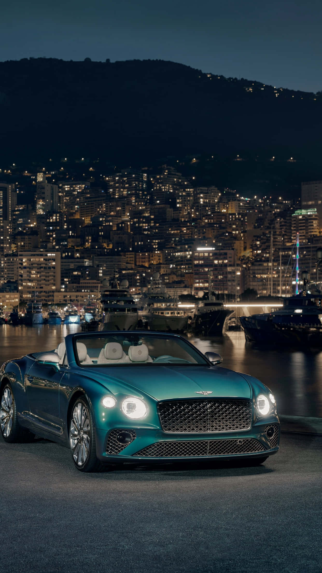 Bentley Continental Gt 桌布 壁紙 — 一架青綠色賓利開篷車在夜燈下閃閃發光，停泊在港口旁，背後的山丘上閃爍著城市景觀。