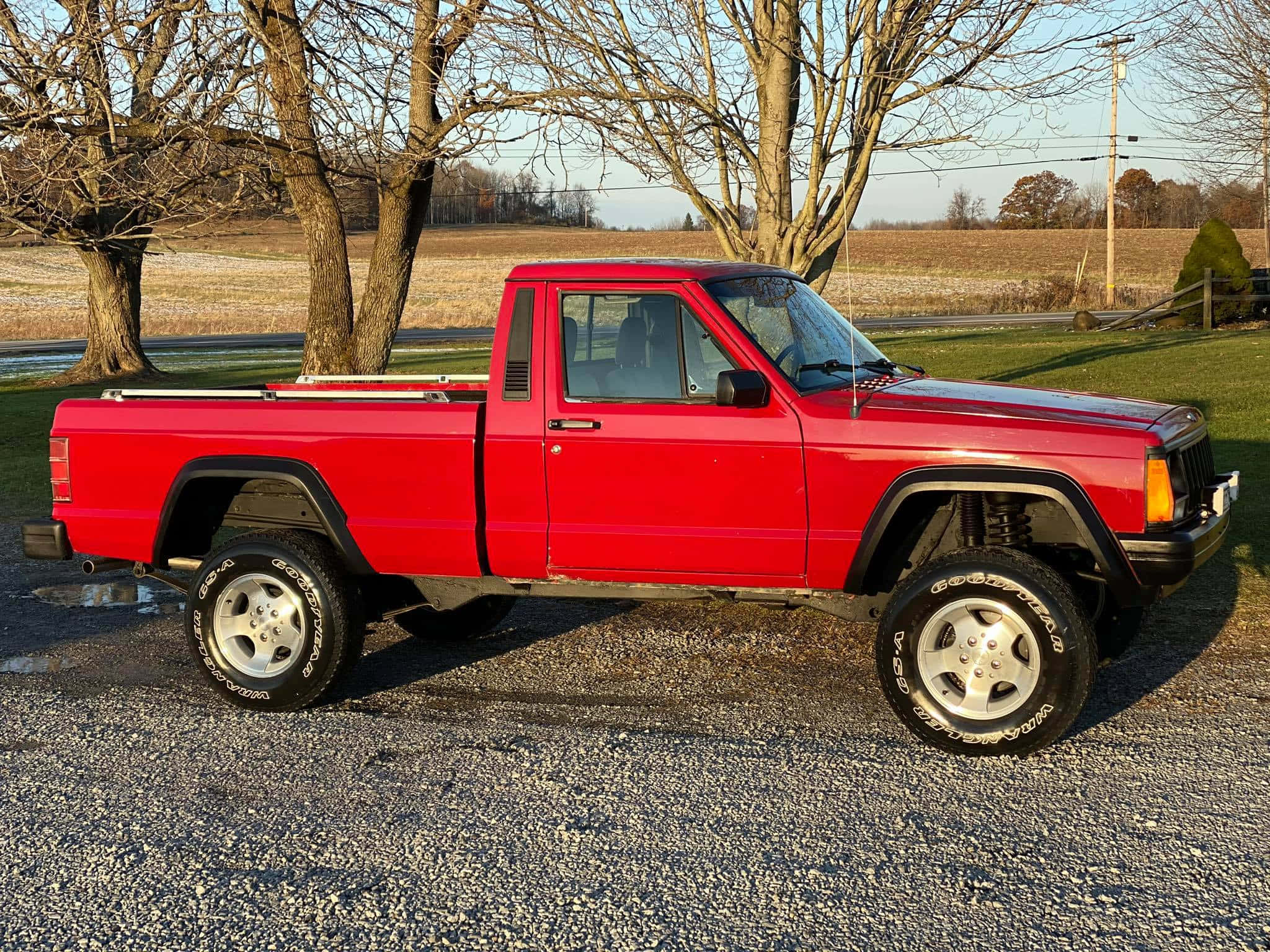 Jeep Comanche 桌布 壁紙 — 一架鮮紅色升高嘅皮卡車停泊喺碎石上，背景係光禿禿嘅樹木同開闊田野，天空晴朗。