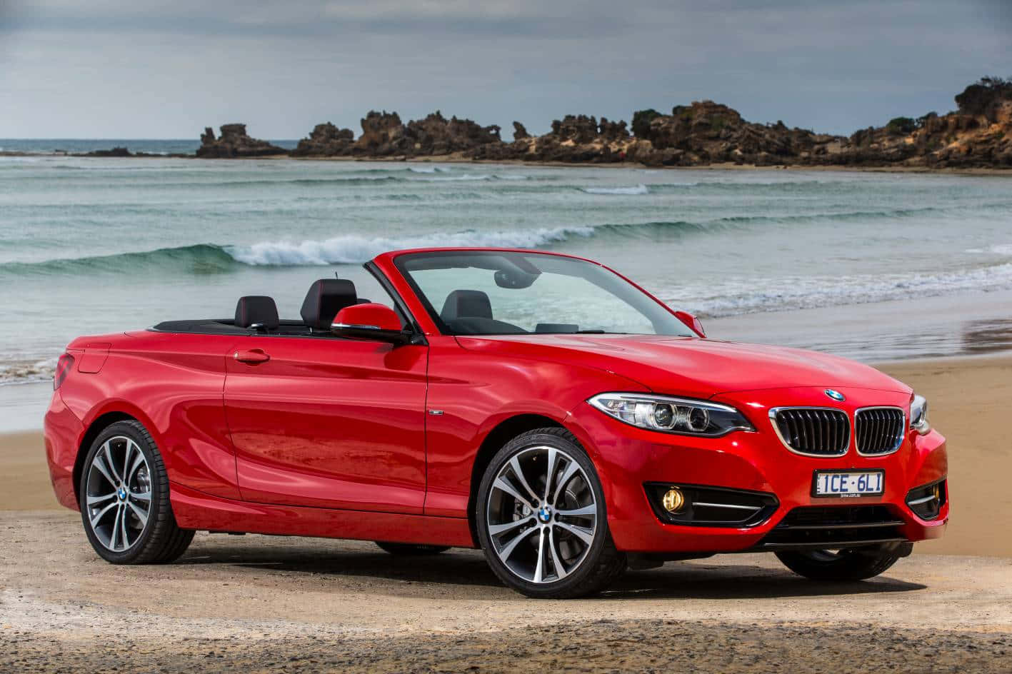 Bmw 2 Series 桌布 壁紙 — 一架鮮紅色寶馬開篷車停泊在沙灘上，背景有海浪和岩石海岬。