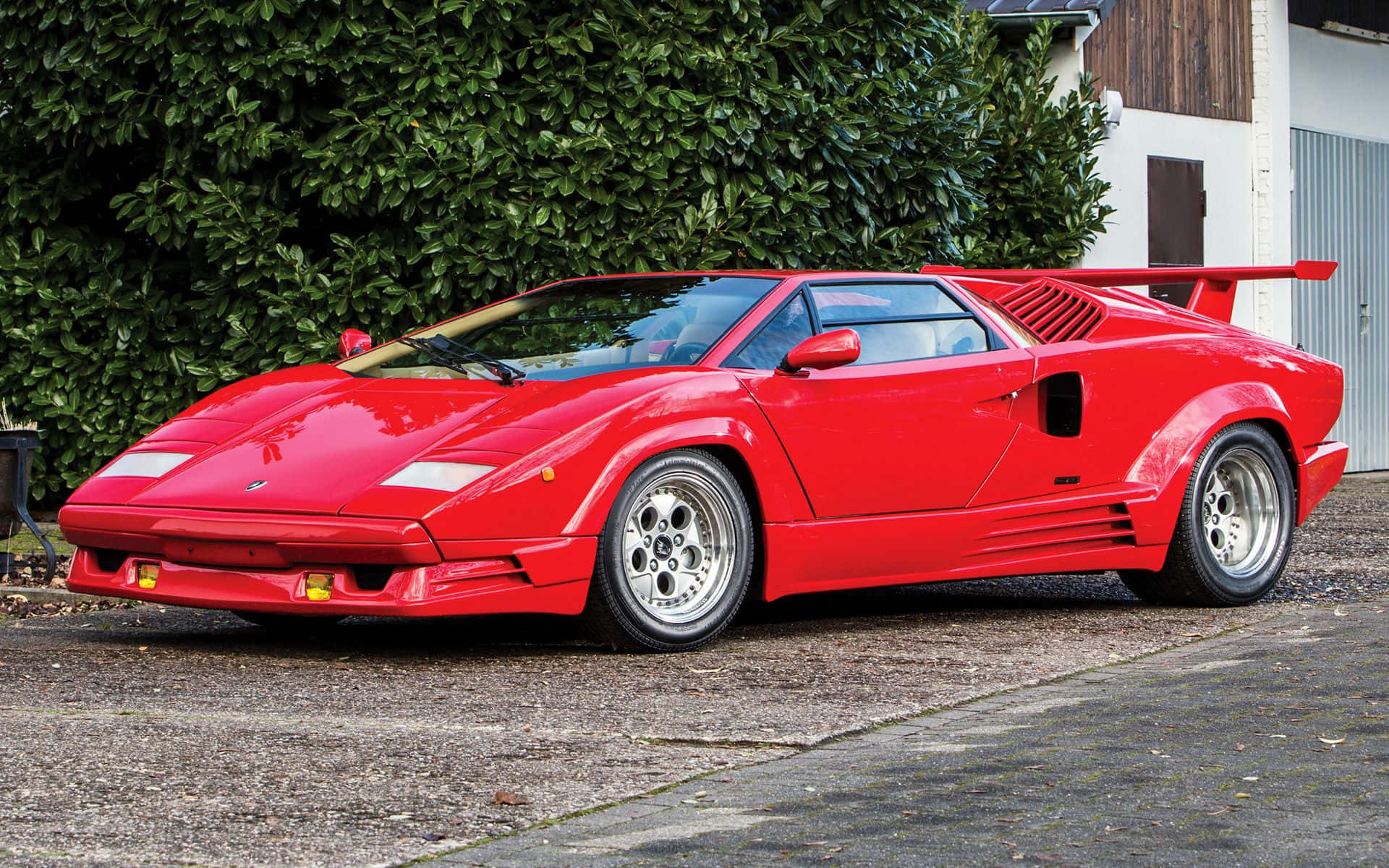 Lamborghini Countach 桌布 壁紙 — 一架鮮紅色林寶堅尼Countach停泊喺碎石車道旁邊，旁邊有茂密嘅綠色籬笆同白色建築物。