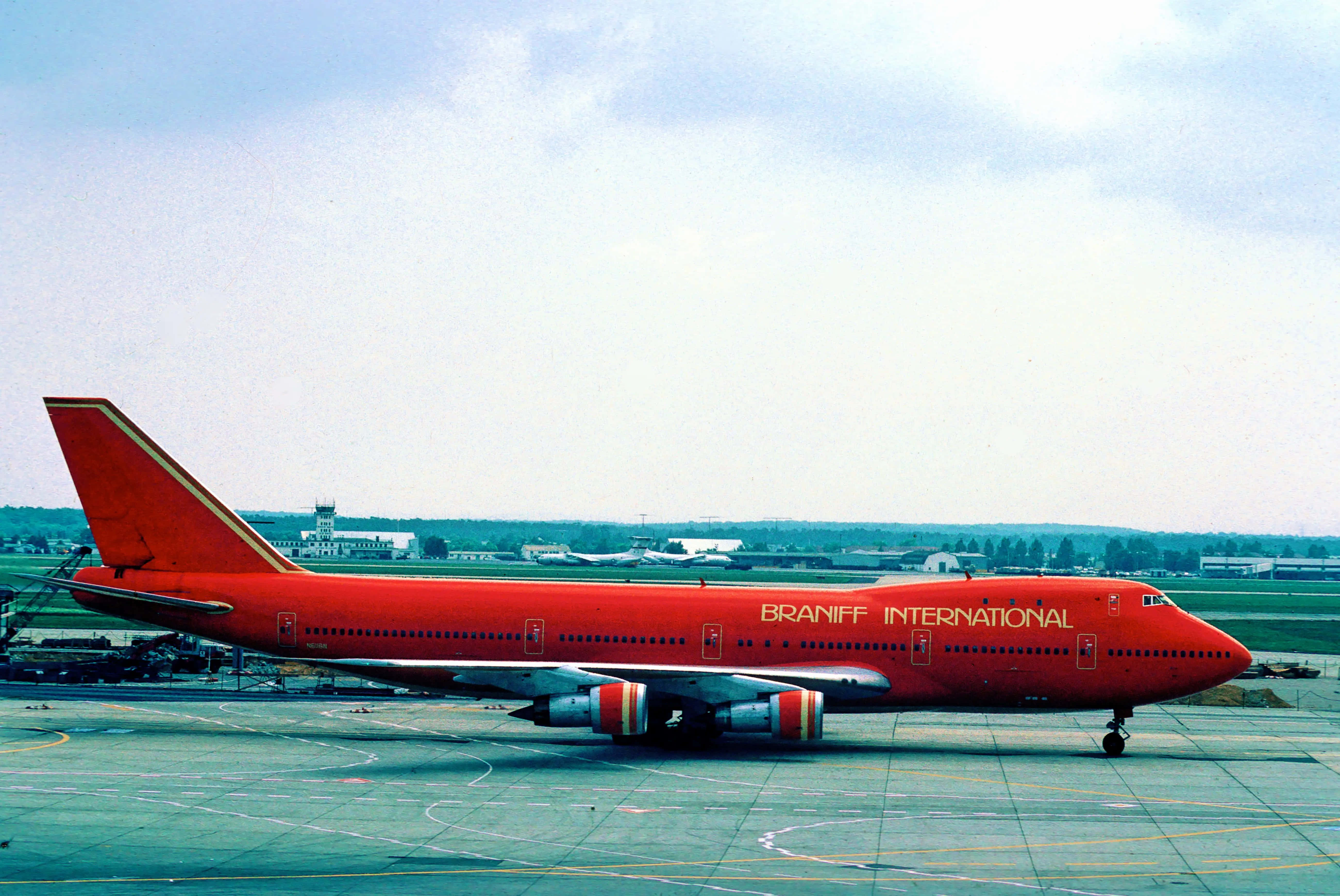 Jumbo Jets 桌布 壁紙 — 一架鮮紅色的 Braniff International 波音 747 停泊在機場停機坪上，天空多雲，遠處有機庫和跑道標記。