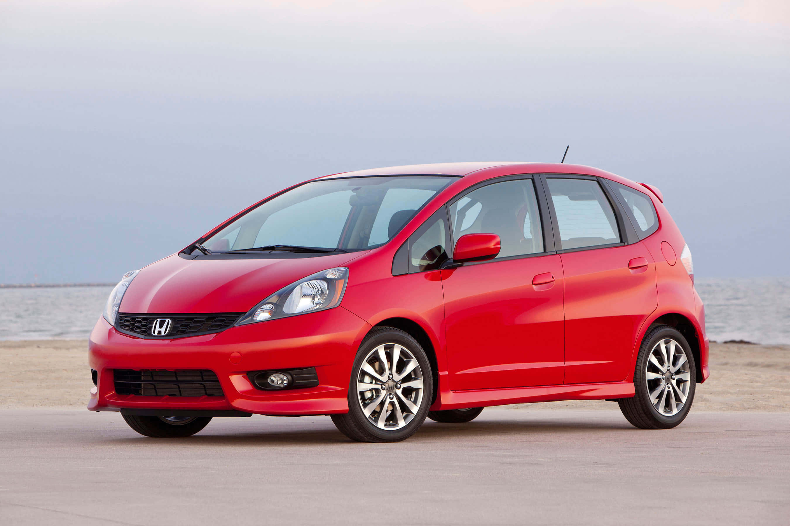 Honda Fit 桌布 壁紙 — 一架鮮紅色的本田Fit停泊在沙灘上，背景是海洋和多雲的天空。