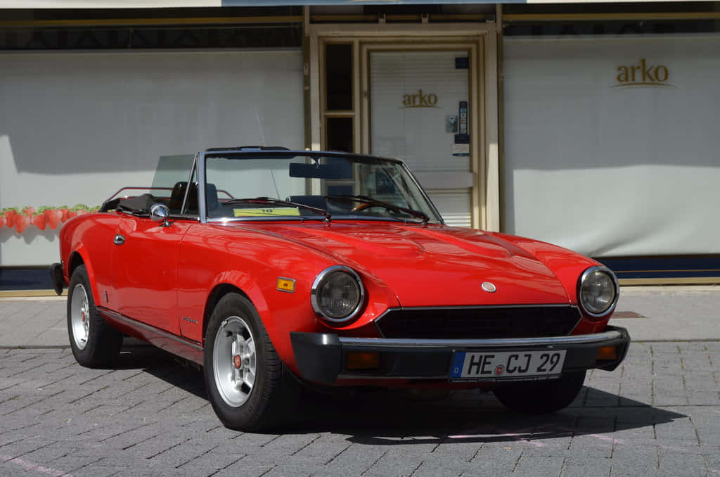 Fiat 124 Spider 桌布 壁紙 — 一架鮮紅色經典開篷跑車停泊在陽光普照的鵝卵石街道上，前方是商店門面。
