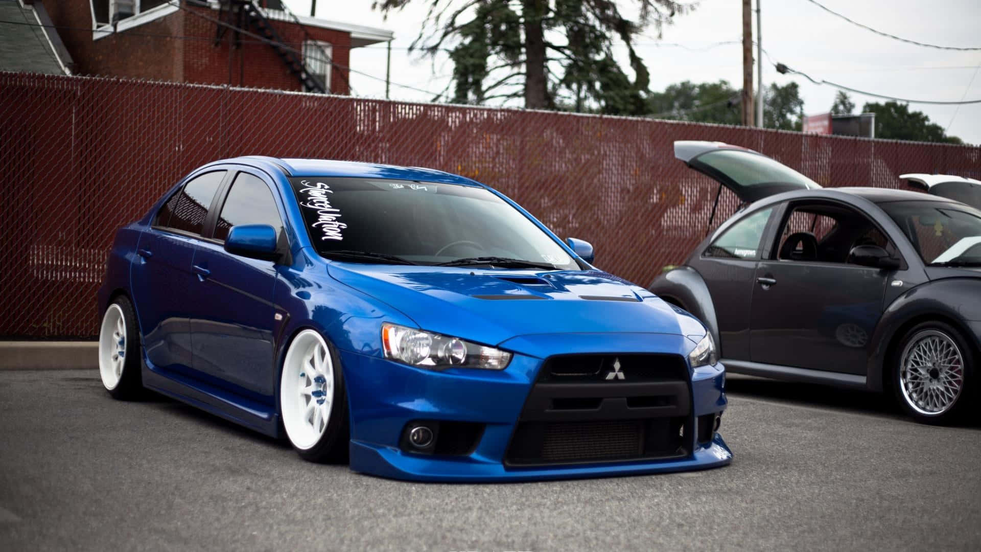 JDM 車 桌布 壁紙 — 一架鮮藍色改裝三菱Lancer Evolution停泊在市區空地，旁邊有一輛打開引擎蓋的灰色福士甲蟲車。