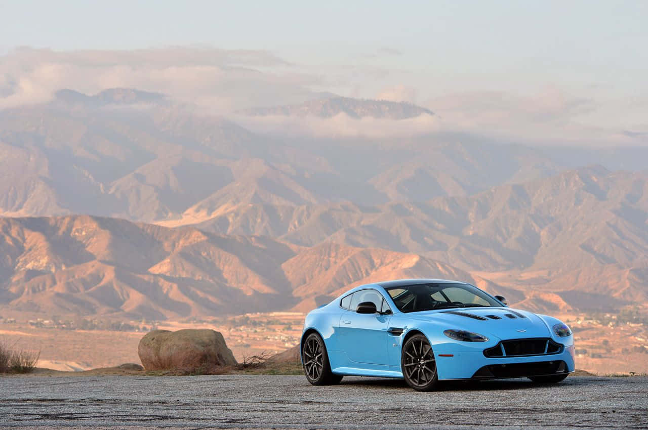 Aston Martin V12 Vantage 桌布 壁紙 — 一架鮮藍色跑車停泊在沙漠觀景點，層層山脈在柔和日光下逐漸消失在霧靄中。
