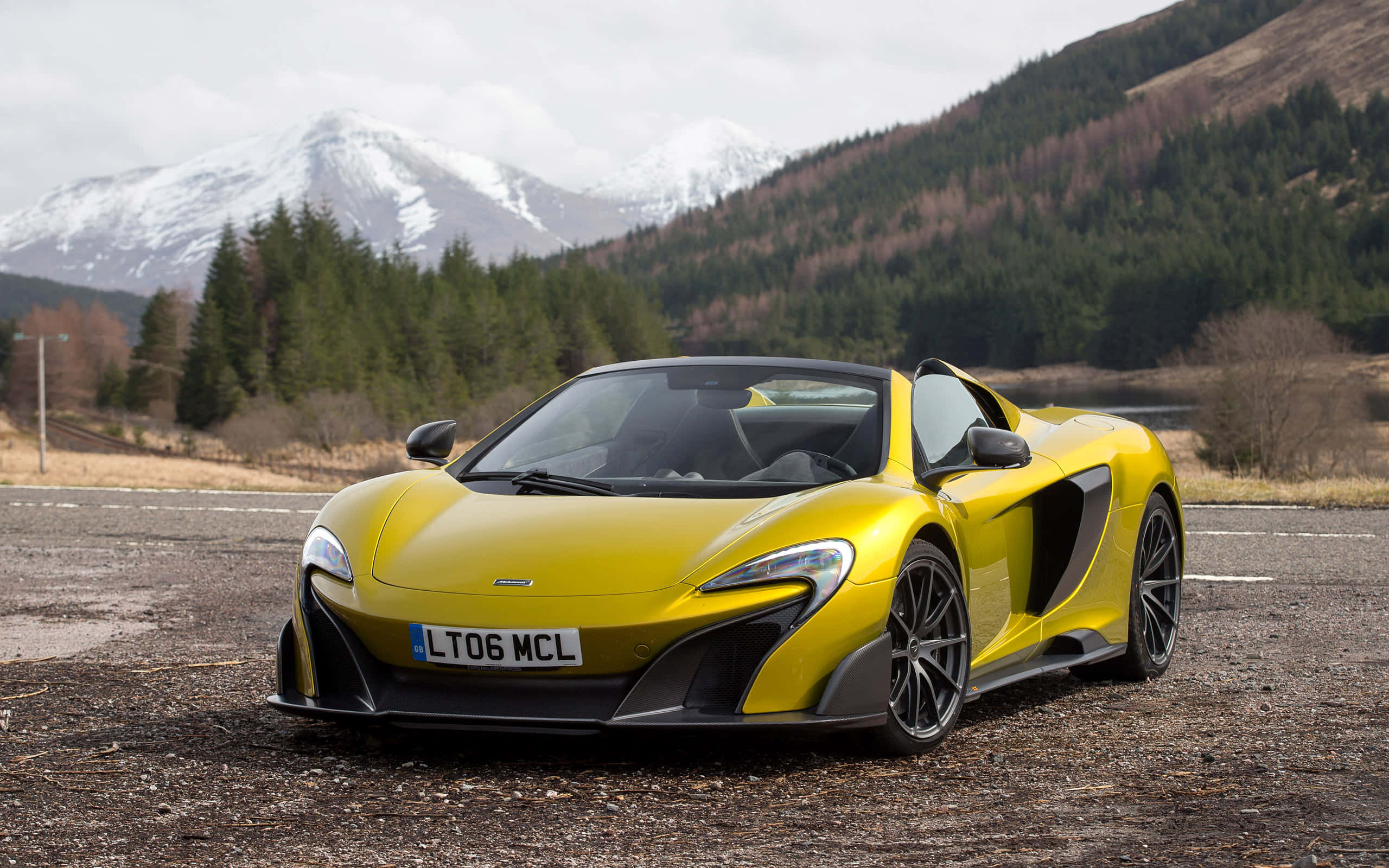 Mclaren 675lt Spider 桌布 壁紙 — 一架鮮黃色McLaren跑車停泊在碎石上，背景是白雪覆蓋的山脈和常綠森林。