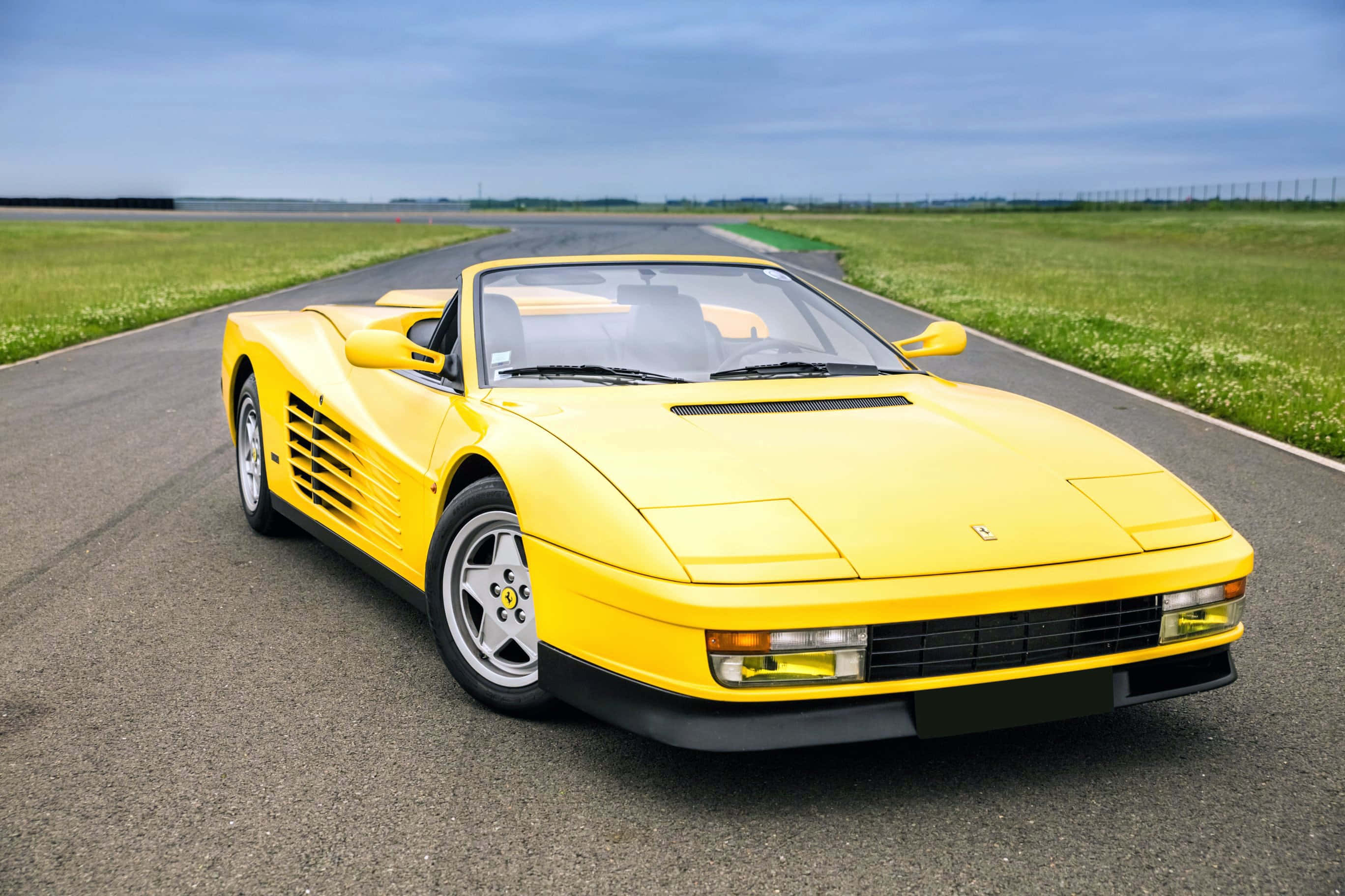 Ferrari Testarossa 桌布 壁紙 — 一架鮮黃色法拉利Testarossa開篷跑車停泊在空曠的瀝青路上，兩旁是綠草，天空多雲。