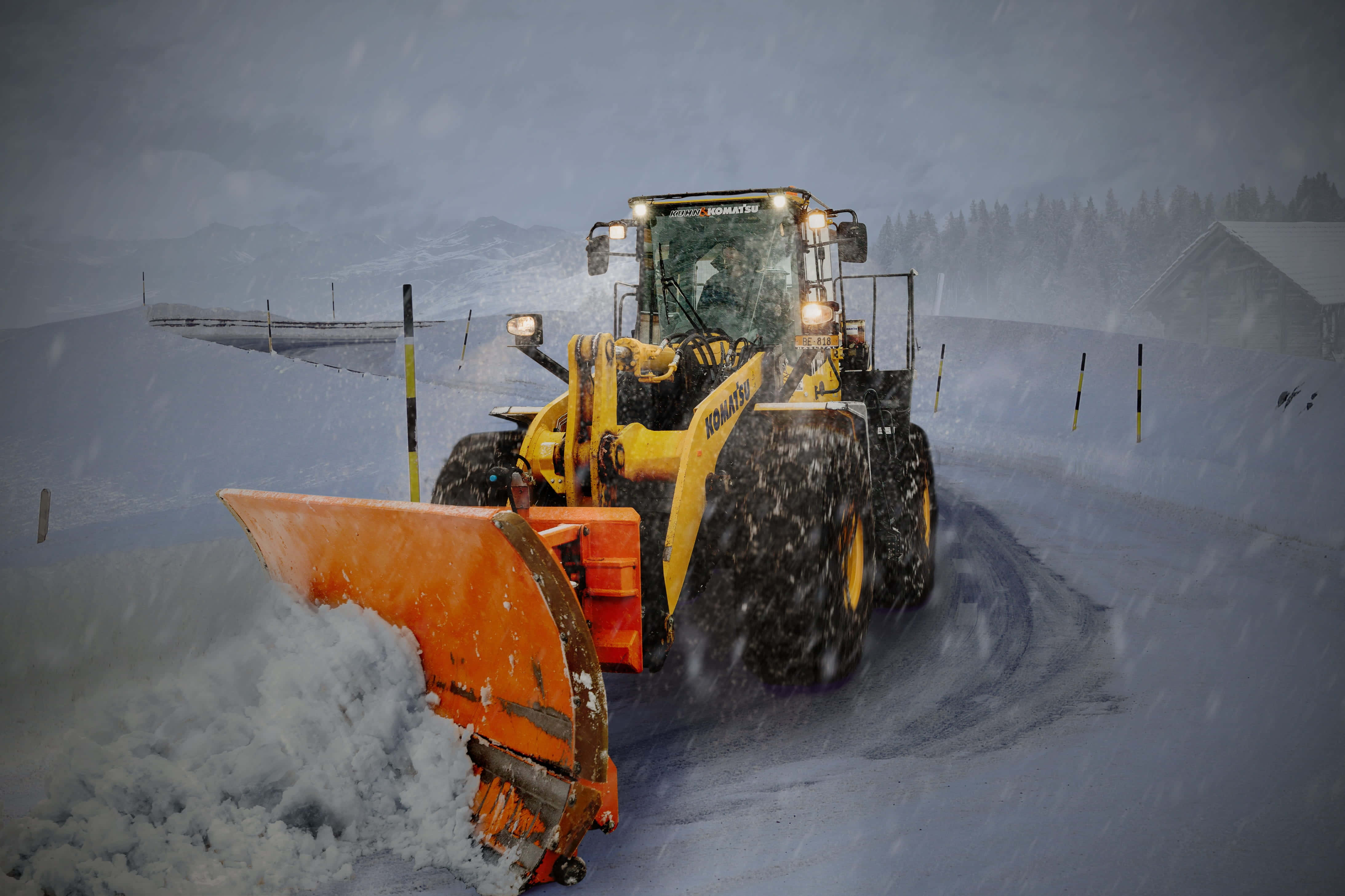 Snowplow 桌布 壁紙 — 一架黃色剷雪車在暴雪中清理積雪道路，背景有山脈和小屋。