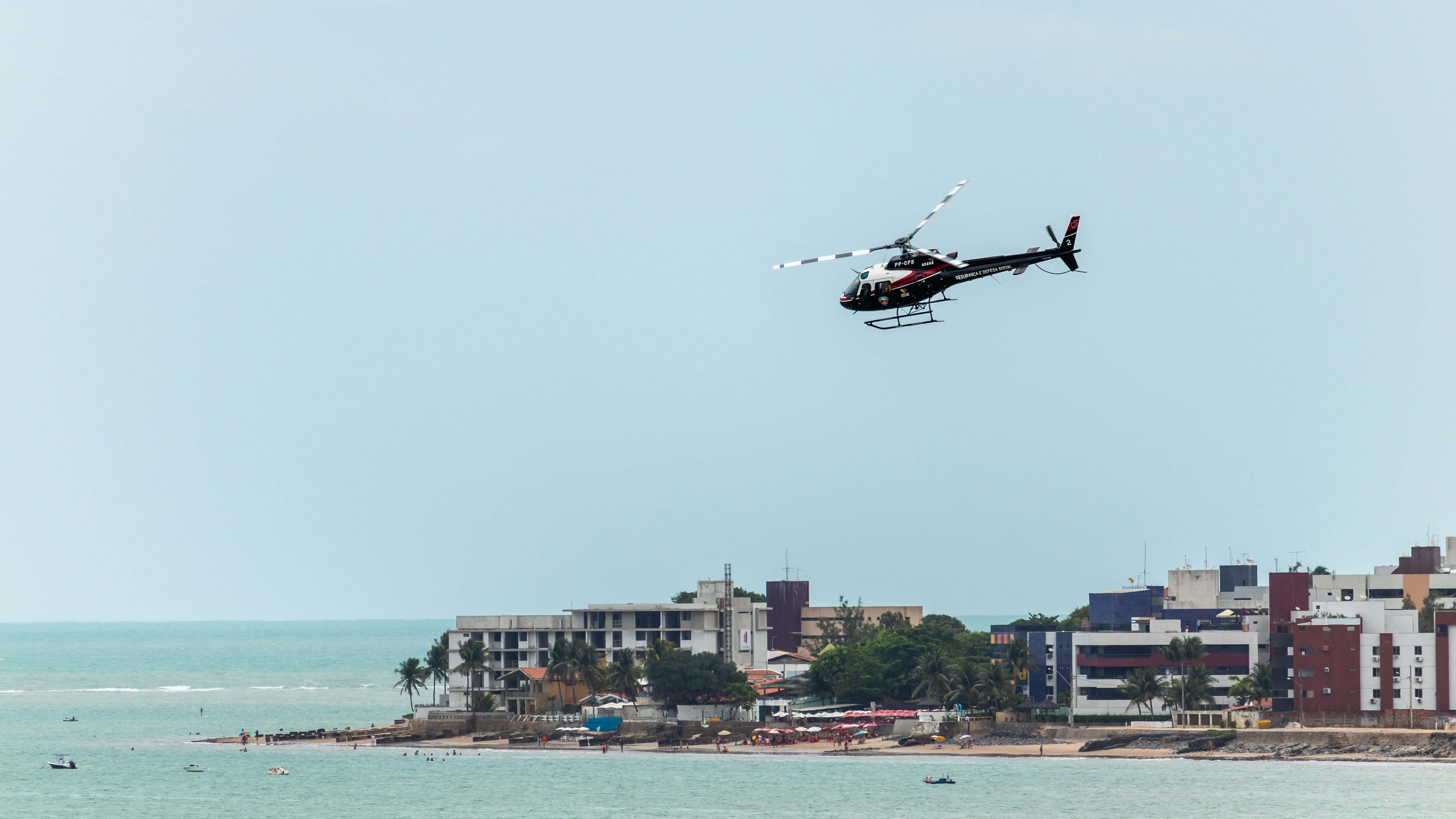 Helicopter 桌布 壁紙 — 一架黑紅色的直升機低飛過一個沿海小鎮，下方有碧綠色的海洋和海濱建築，天空朦朧。