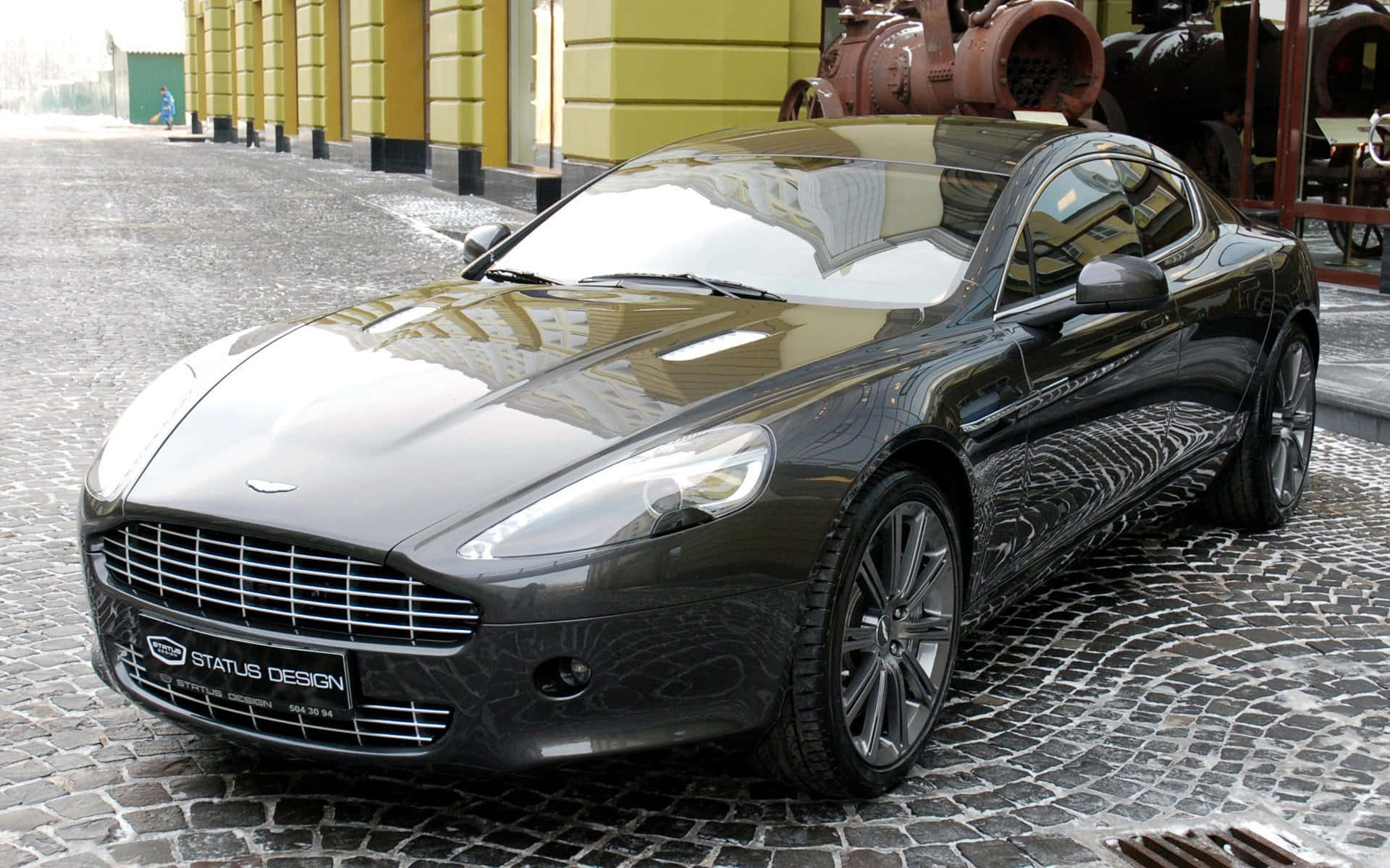 Aston Martin Rapide S 桌布 壁紙 — 一架黑色Aston Martin跑車在濕滑的鵝卵石街道上閃閃發光，旁邊是一座黃色建築物。