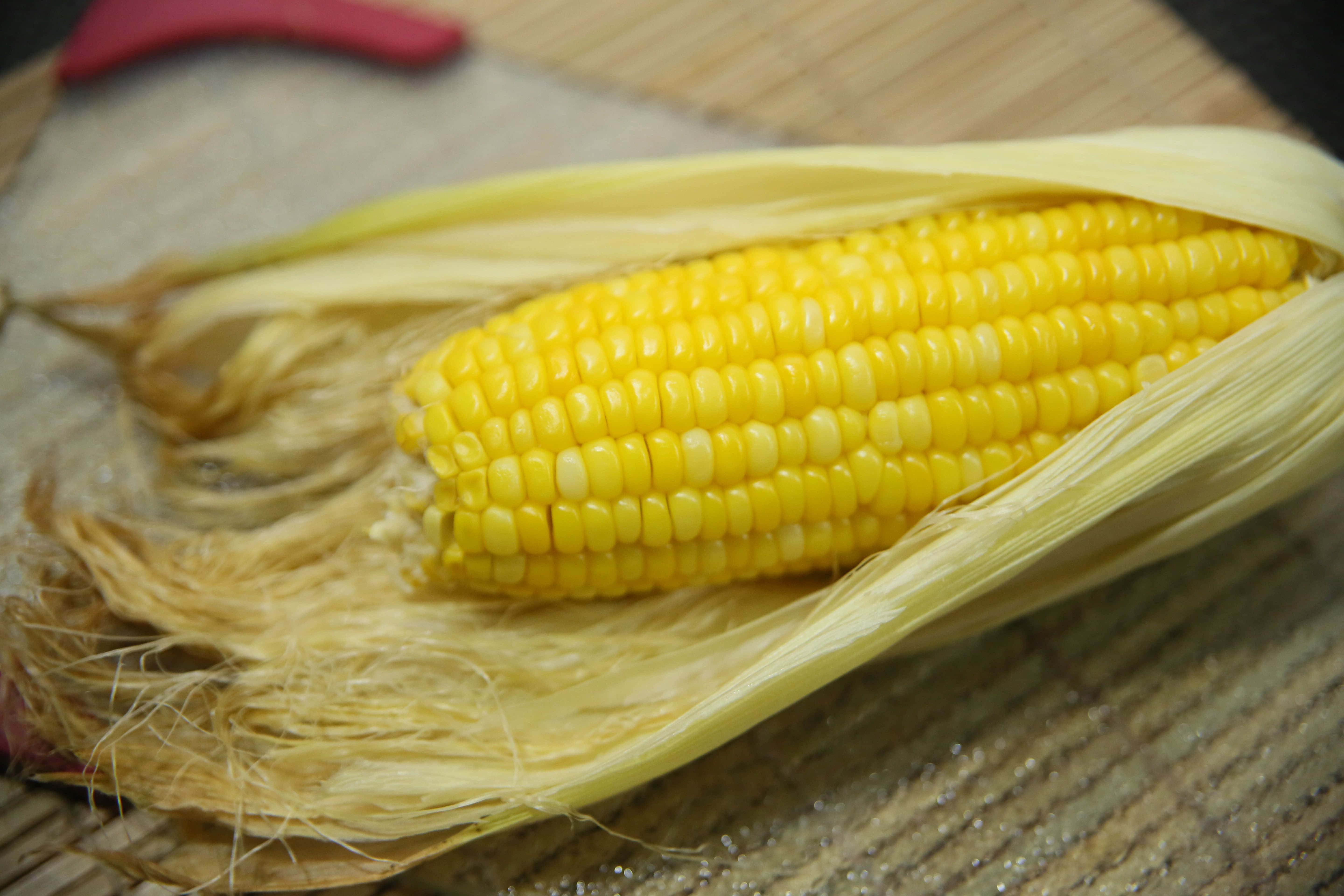 Corn 桌布 壁紙 — 一根剛剝殼的亮黃色玉米穗放在編織竹蓆上，附近有一個紅色手柄的工具。