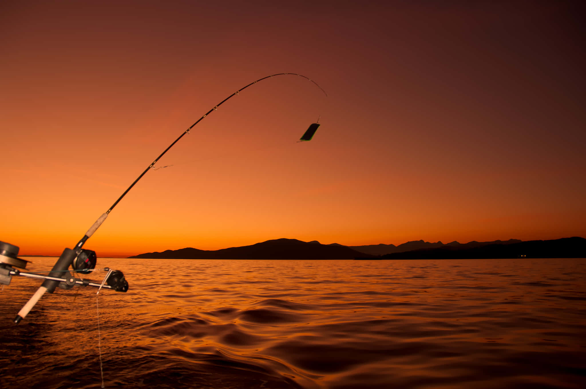 Fishing Rod 桌布 壁紙 — 一根魚竿在平靜的水面和遠山之上，對著火紅的橙色夕陽戲劇性地彎曲。