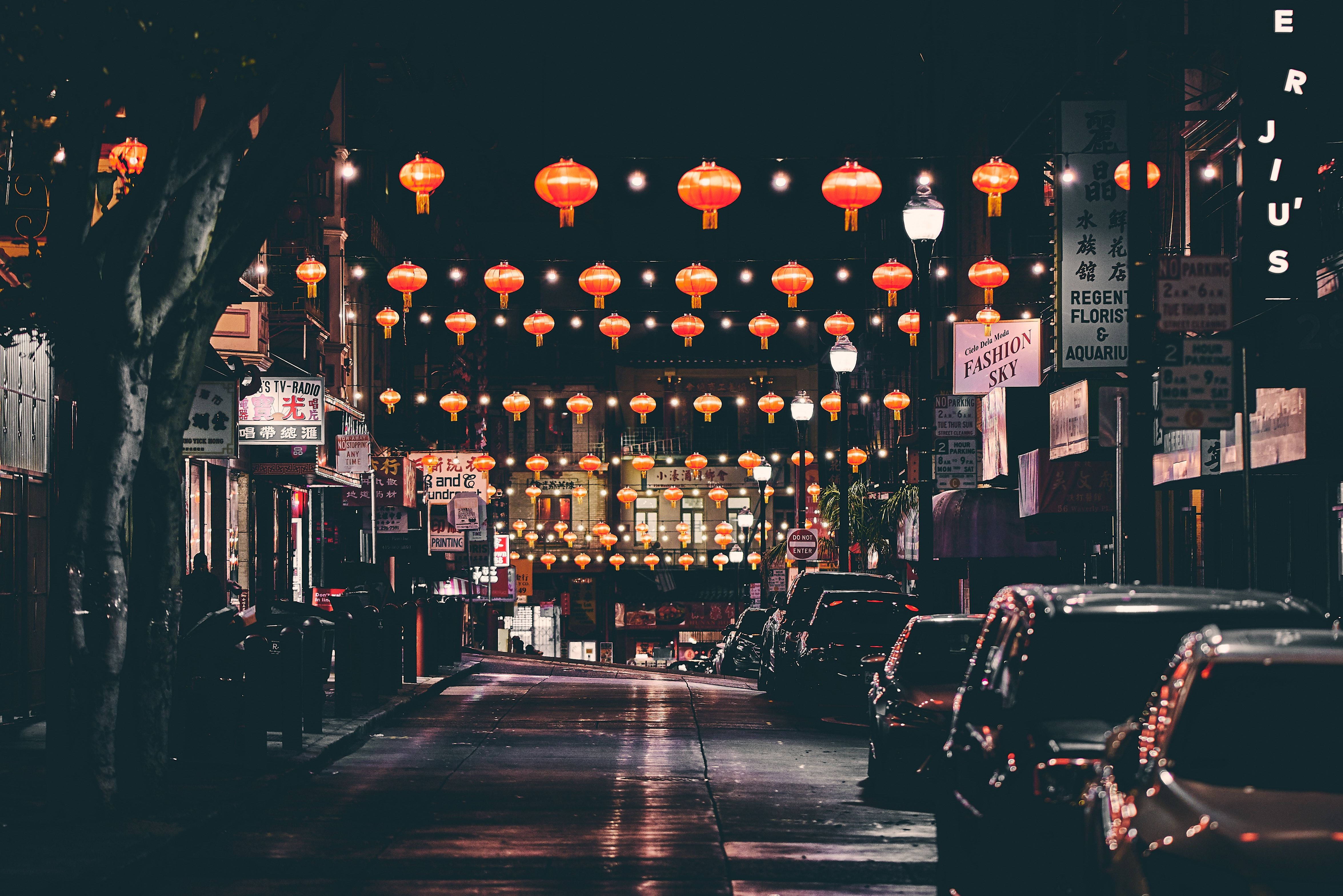 三藩市 桌布 壁紙 — 一條充滿活力的夜晚街道，裝飾著懸掛在停泊汽車和明亮店面上方的發光紅燈籠。