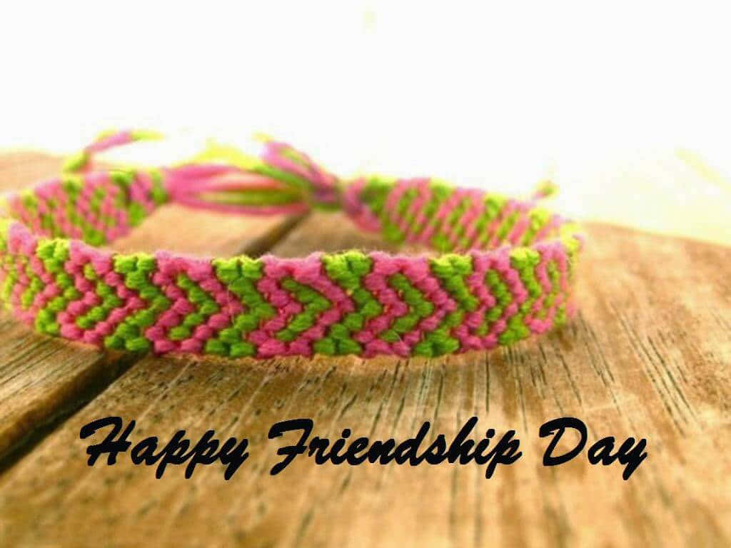 Friendship Day 桌布 壁紙 — 一條充滿活力的粉紅和綠色編織友誼手鍊放在質樸的木質表面上，帶有'Happy Friendship Day'文字。