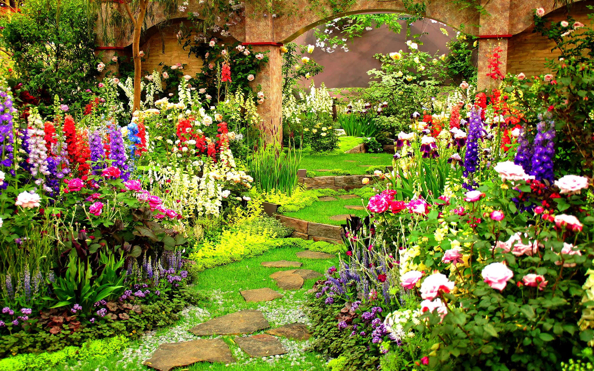 Garden Photography 桌布 壁紙 — 一條充滿活力的花園小徑，兩旁種滿色彩繽紛的花朵和石板，通向一個質樸的拱形棚架下。