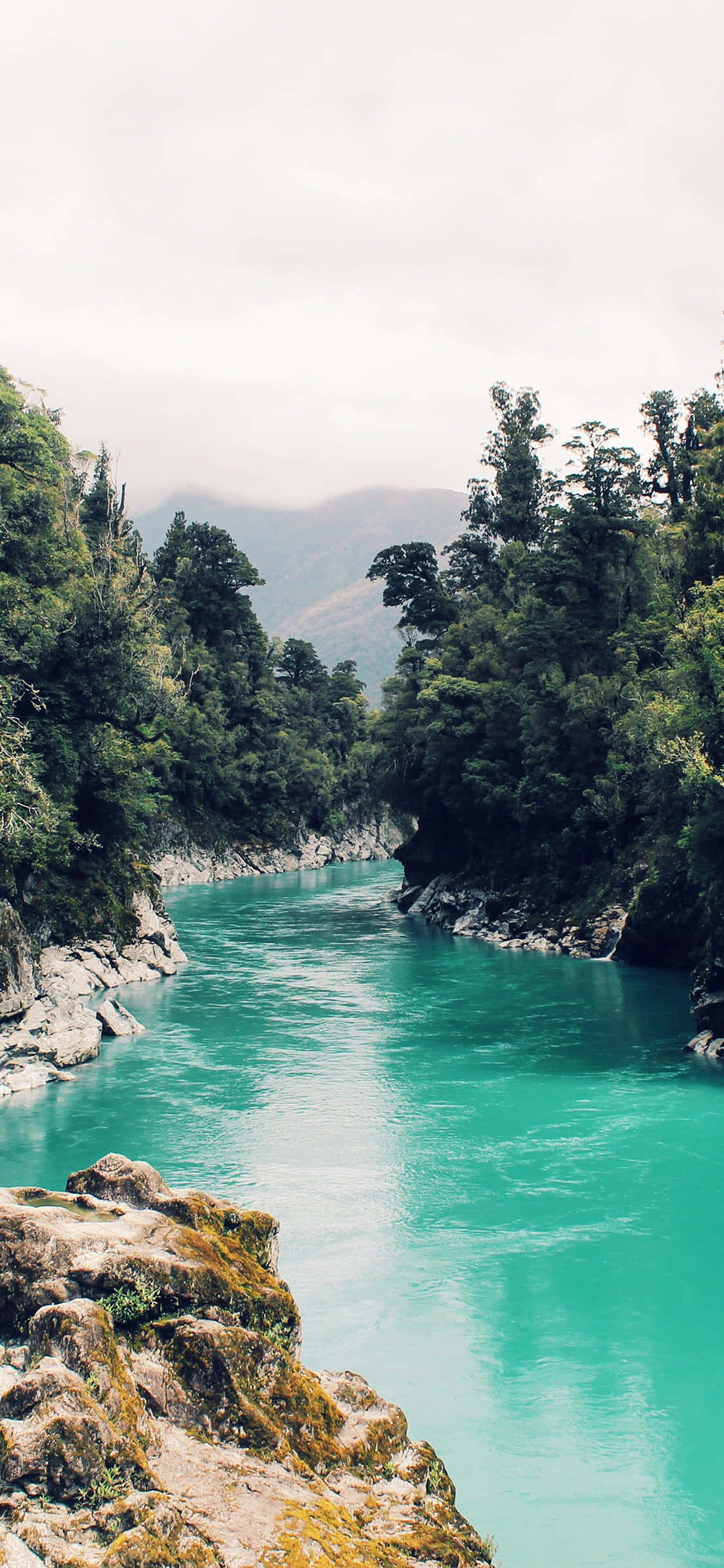 iPhone X Nature 桌布 壁紙 — 一條充滿活力的藍綠色河流蜿蜒穿過鬱鬱蔥蔥的綠色森林峽谷，天空多雲。