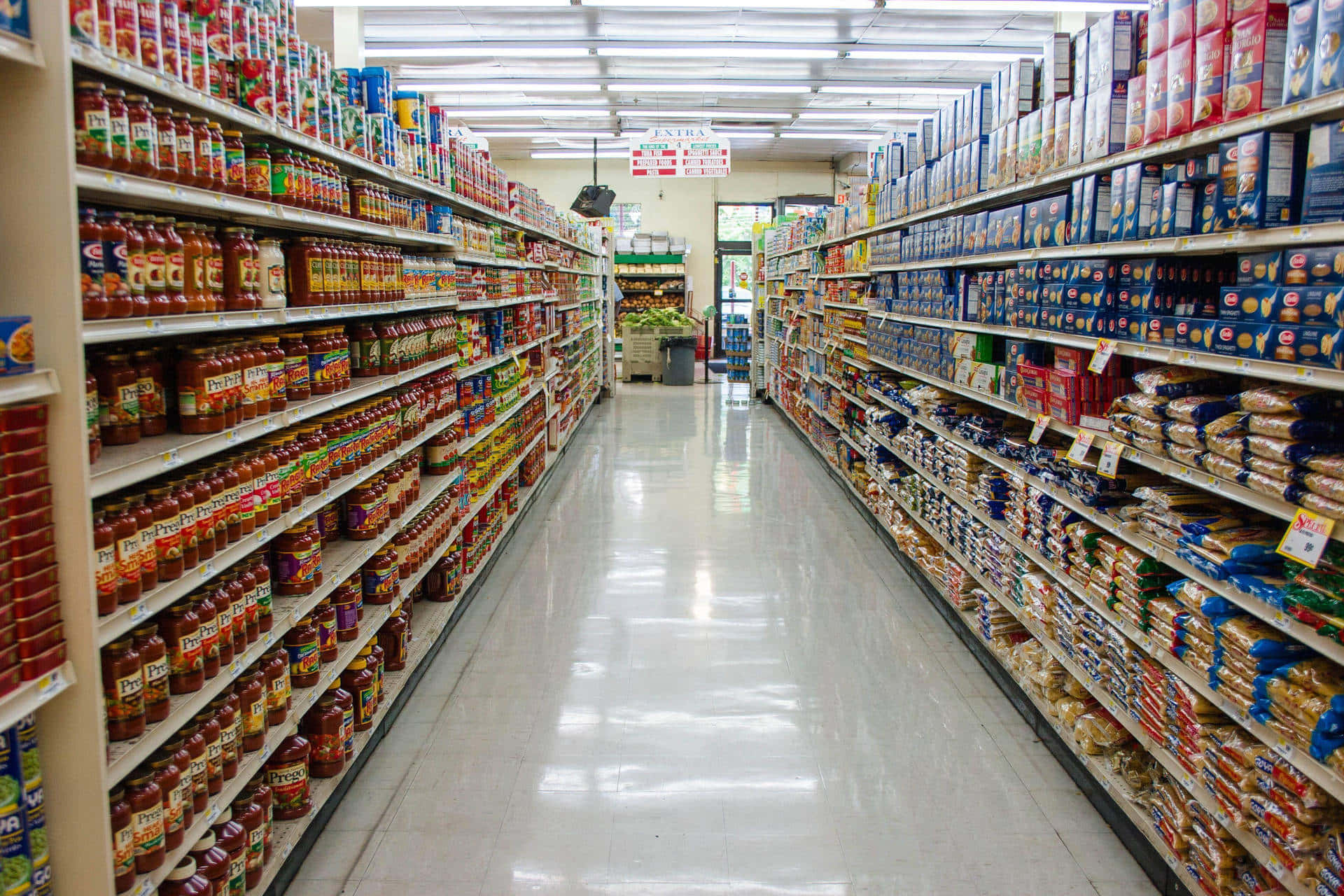 Grocery Store 桌布 壁紙 — 一條光線充足嘅長超市通道，兩旁擺滿意粉醬同麵包產品嘅貨架。