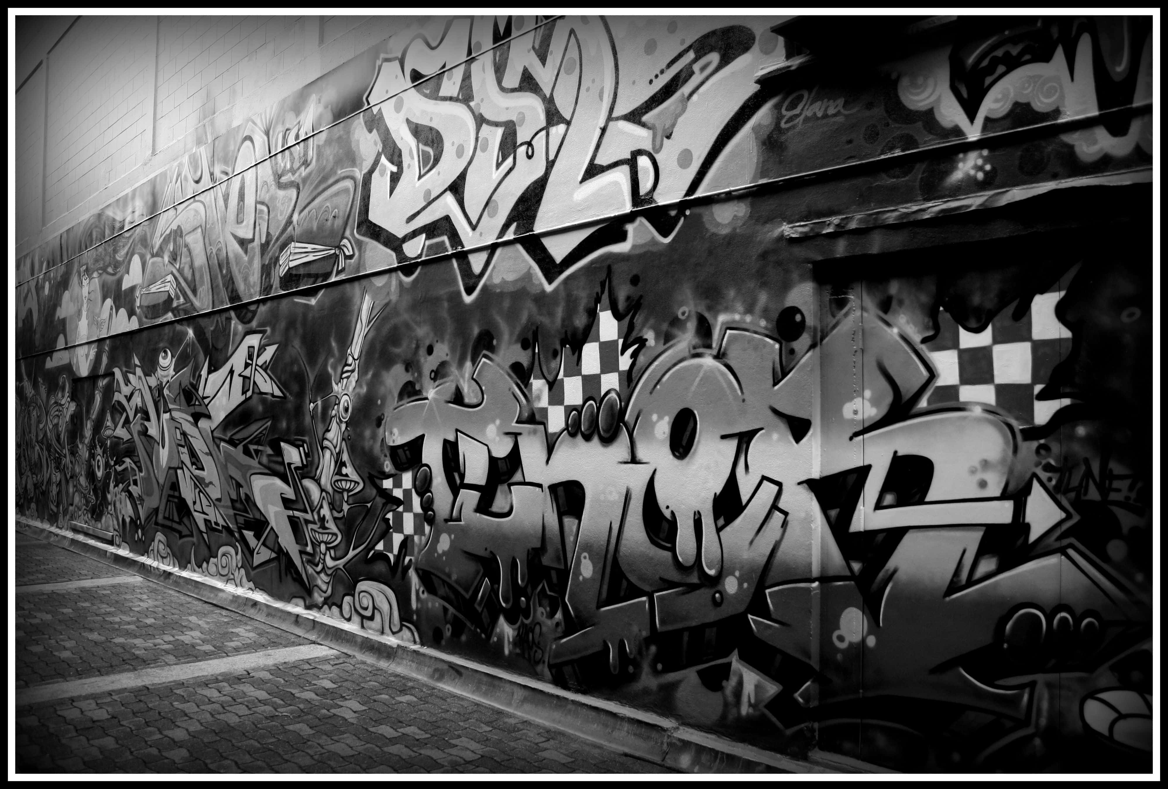 Black And White Graffiti 桌布 壁紙 — 一條單色城市小巷的牆壁，上面覆蓋著大膽的塗鴉標籤和帶有格仔圖案的風格化字體。