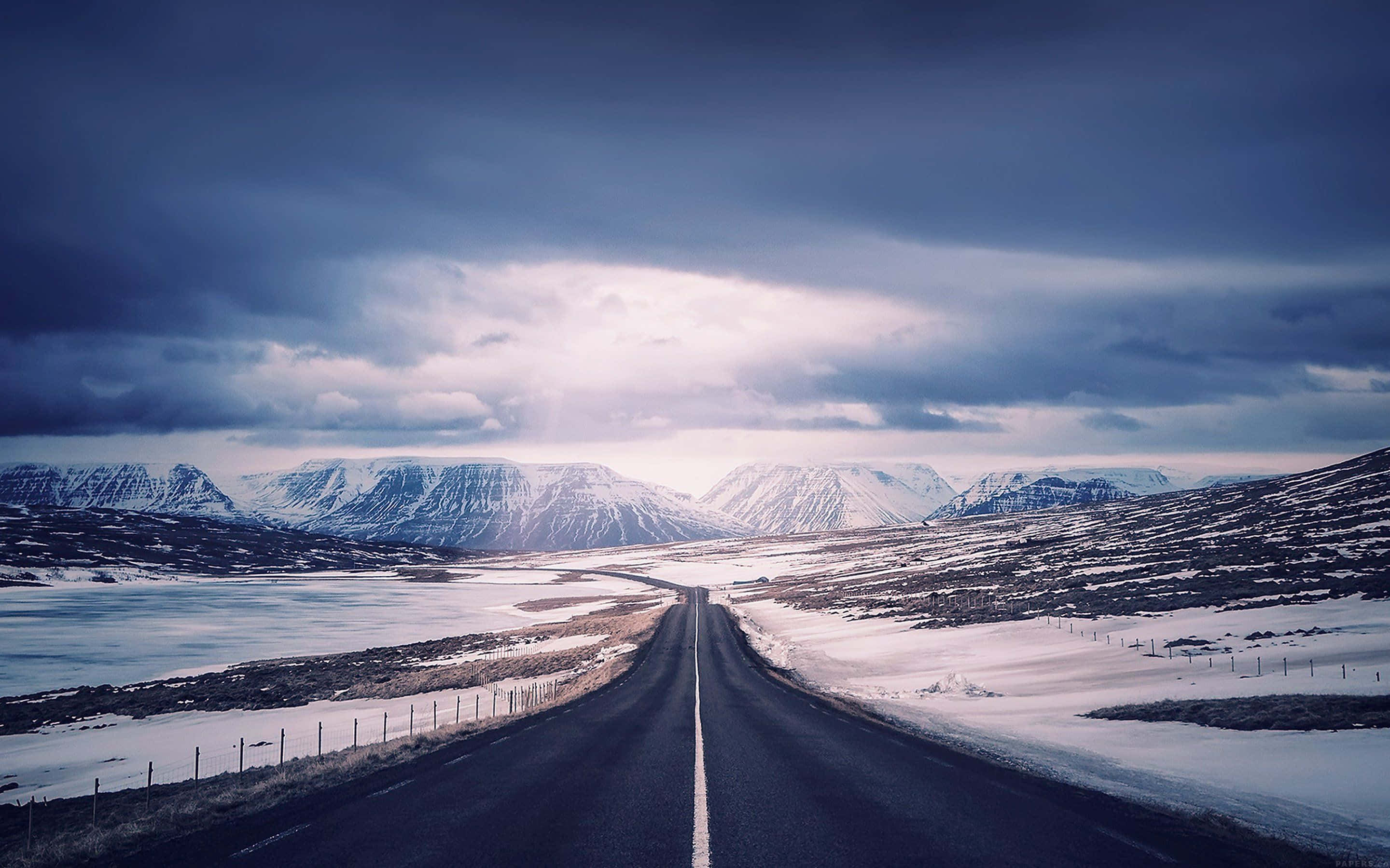 Icy Road 桌布 壁紙 — 一條孤獨的瀝青路在冬季戲劇性的多雲天空下延伸向積雪覆蓋的山脈。