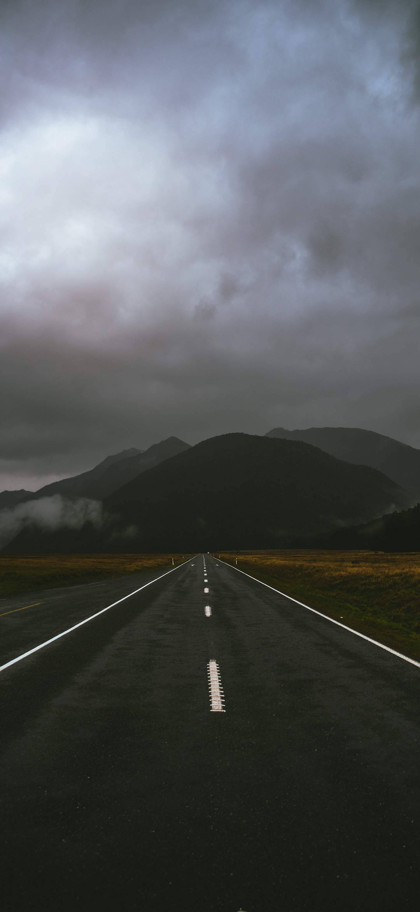 道路 桌布 壁紙 — 一條孤獨的瀝青路在陰沉密佈的天空下延伸向遠山，霧氣纏繞著山坡。