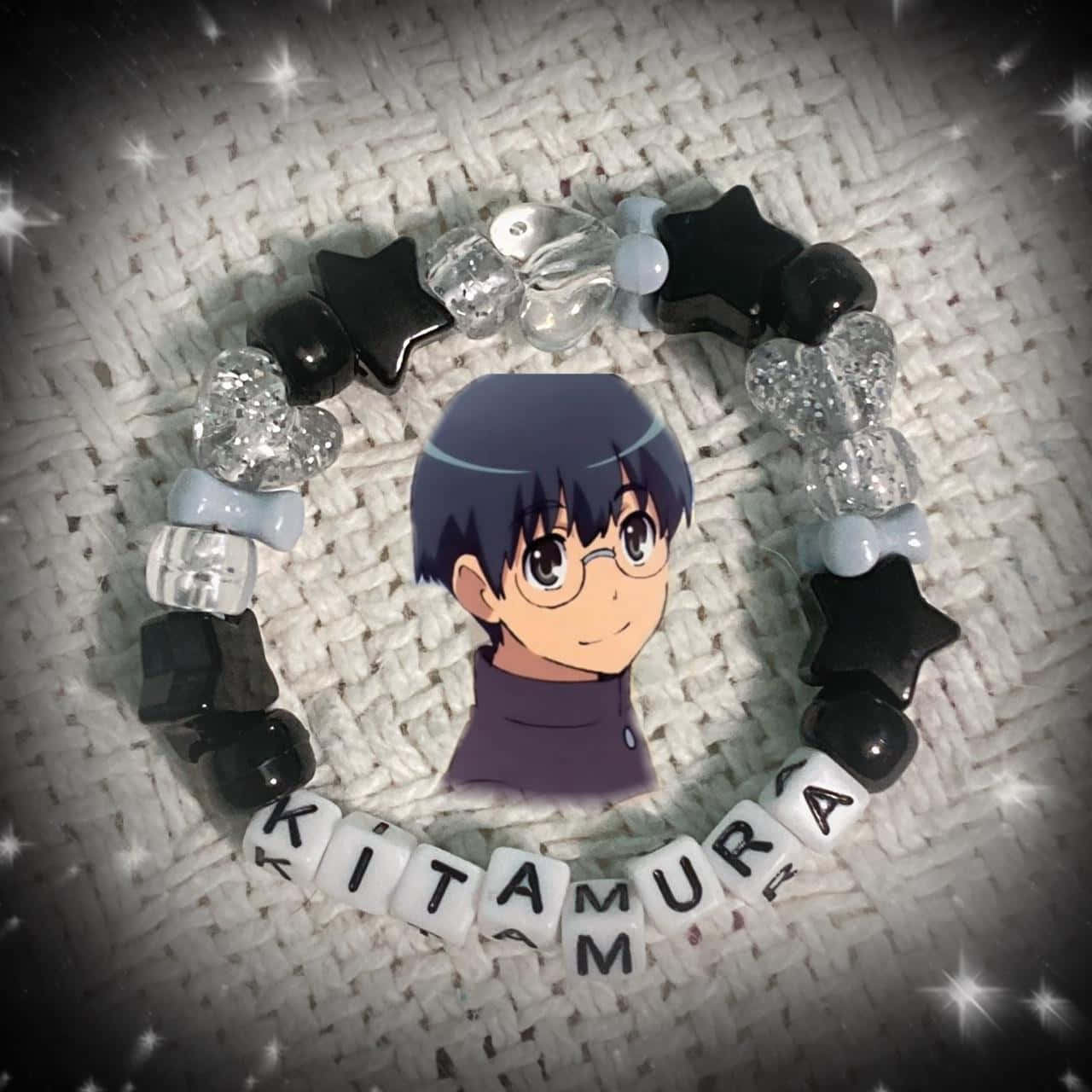 Yusaku Kitamura 桌布 壁紙 — 一條定制珠串手鏈，用黑色星星和閃粉珠子在紋理白色布料背景上拼出Kitamura，圍繞一個印刷的動漫角色肖像。