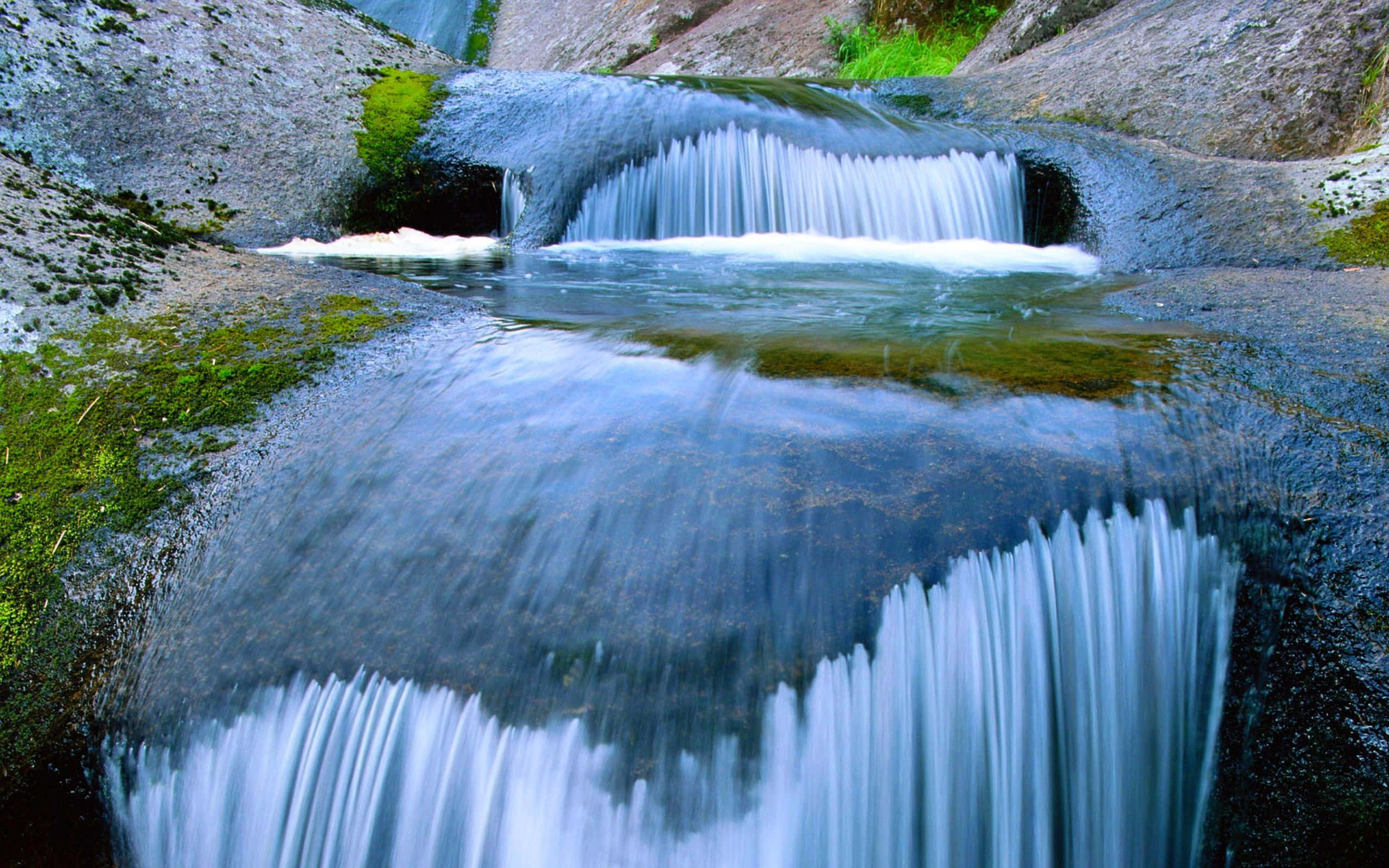 Spring Water 桌布 壁紙 — 一條寧靜的山間溪流在長時間曝光拍攝中流過長滿苔蘚的岩石，營造出絲滑的白色水流，與清涼的藍色和灰色石頭形成對比。