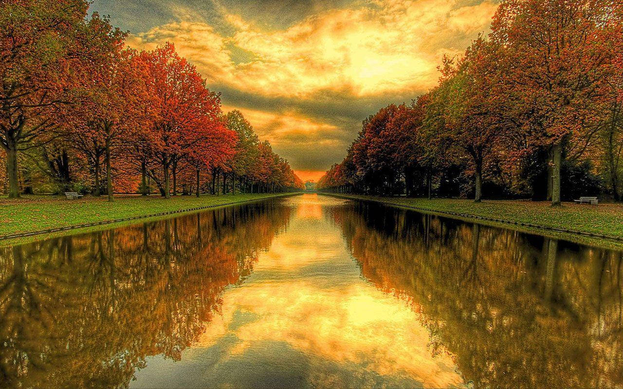 Beautiful Nature Photography 桌布 壁紙 — 一條寧靜的秋天運河，兩旁是火紅和橙色的樹木，在金色夕陽天空下倒映在平靜的水面上。