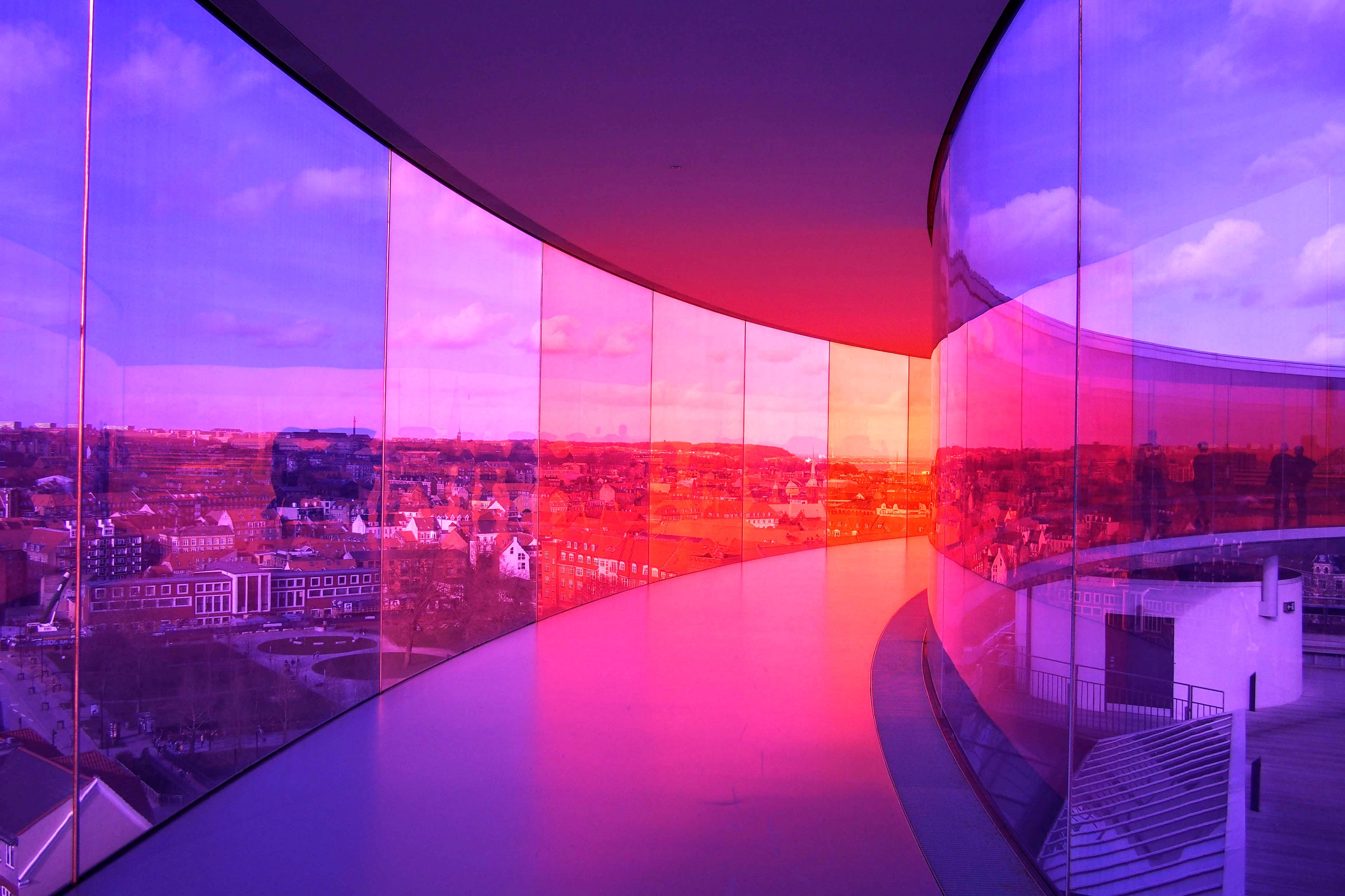 Denmark Panorama Bridge 壁紙 — 一條彎曲的玻璃步道沐浴在充滿活力的粉紅色和紫色燈光中，反射出暮色天空下的城市景觀。