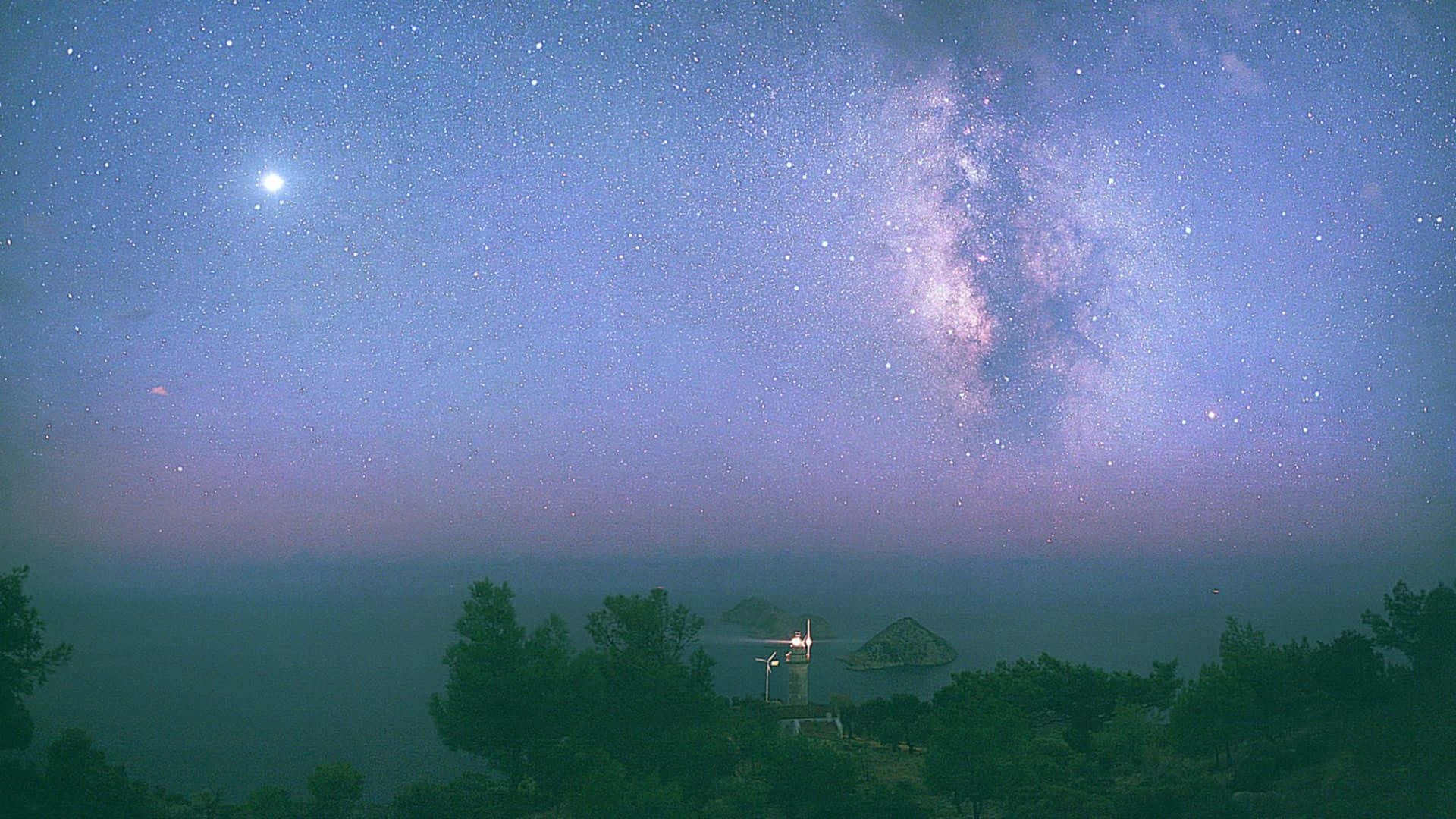 月亮與星星動畫 桌布 壁紙 — 一條明亮的銀河在霧氣瀰漫的海岸景觀上空拱起，有一座發光的燈塔和剪影樹木，背景是星光閃爍的黃昏天空。