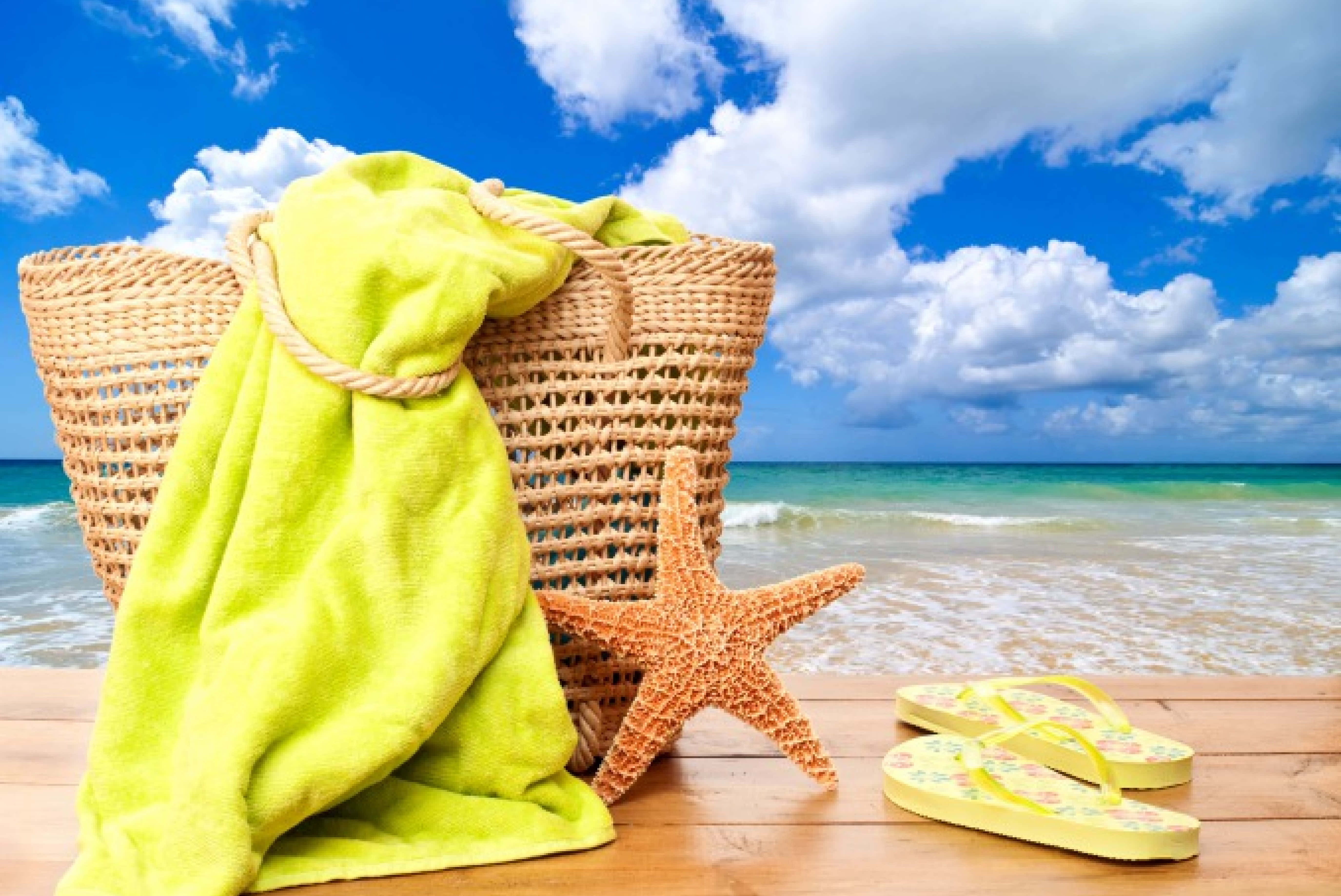 Beach Accessories 桌布 壁紙 — 一條明亮的青檸色毛巾搭在編織沙灘袋上，旁邊有海星和黃色人字拖，放在木製甲板上，俯瞰著碧綠的海浪，天空湛藍，白雲蓬鬆。