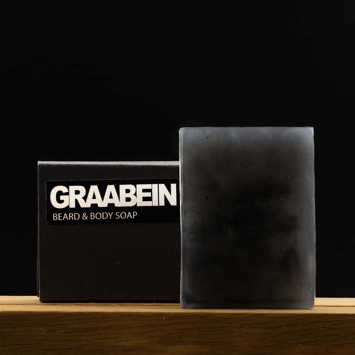 Black Soap 桌布 壁紙 — 一條時尚的黑色Graabein鬍鬚和身體肥皂條，旁邊放著相配的盒子，在溫暖的木架上，背景為深色。