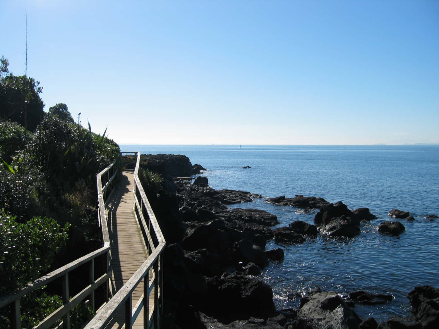 Seaside 桌布 壁紙 — 一條木製步道沿著岩石海岸線蜿蜒，上方是清澈的藍天，海面平靜。