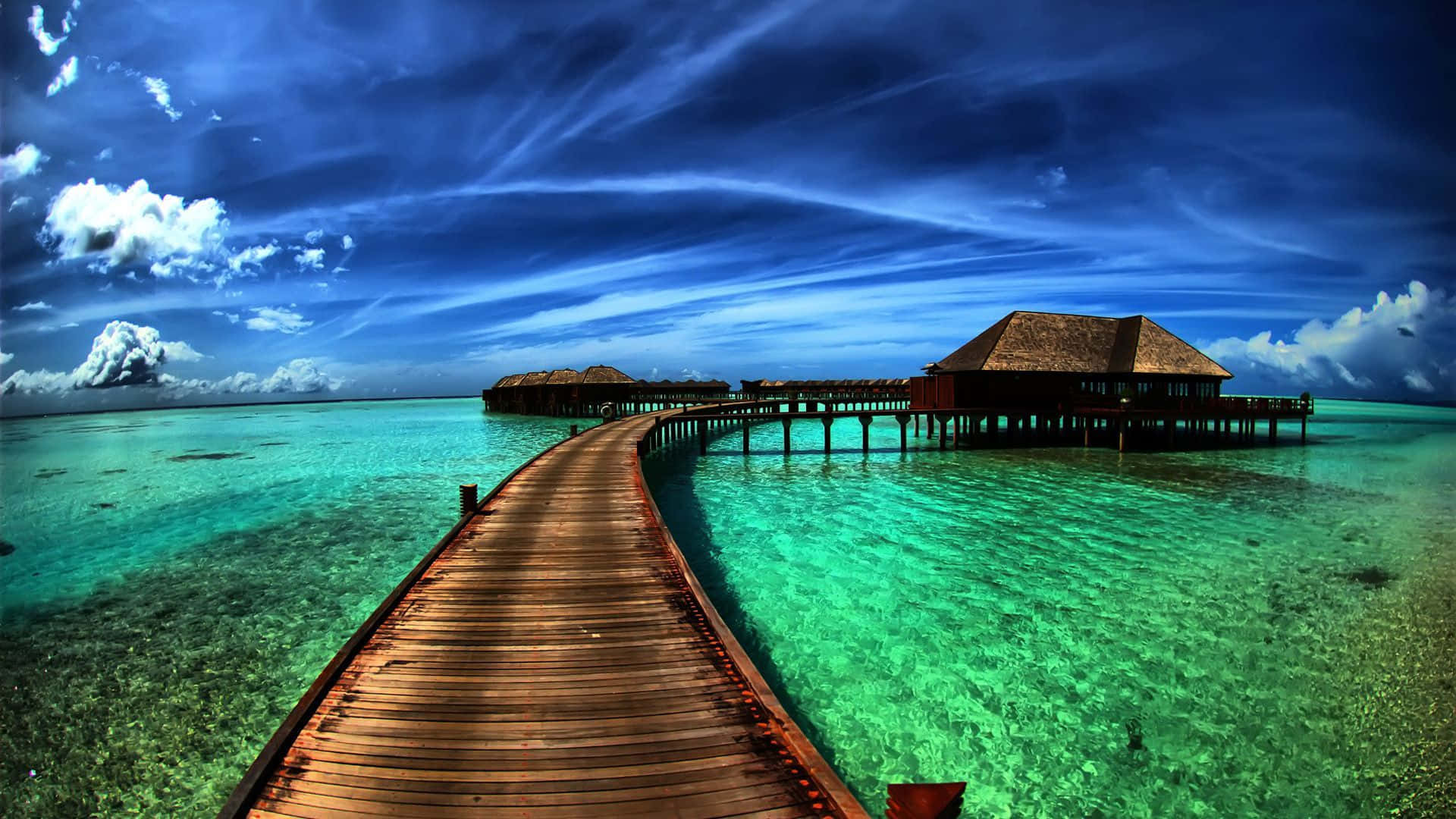 Maldives Beach 桌布 壁紙 — 一條木製碼頭延伸在清澈碧綠的海水上，通往水上平房，上方是帶有白雲的戲劇性藍天。