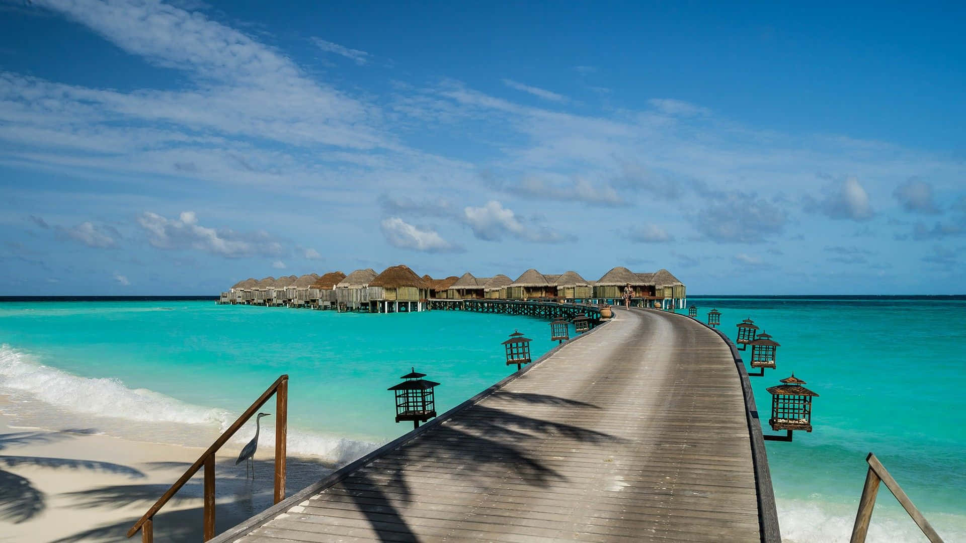 Maldives Beach 桌布 壁紙 — 一條木製碼頭延伸在碧綠色的海水上，通往水上平房，上方是晴朗的藍天，點綴著幾朵白雲。