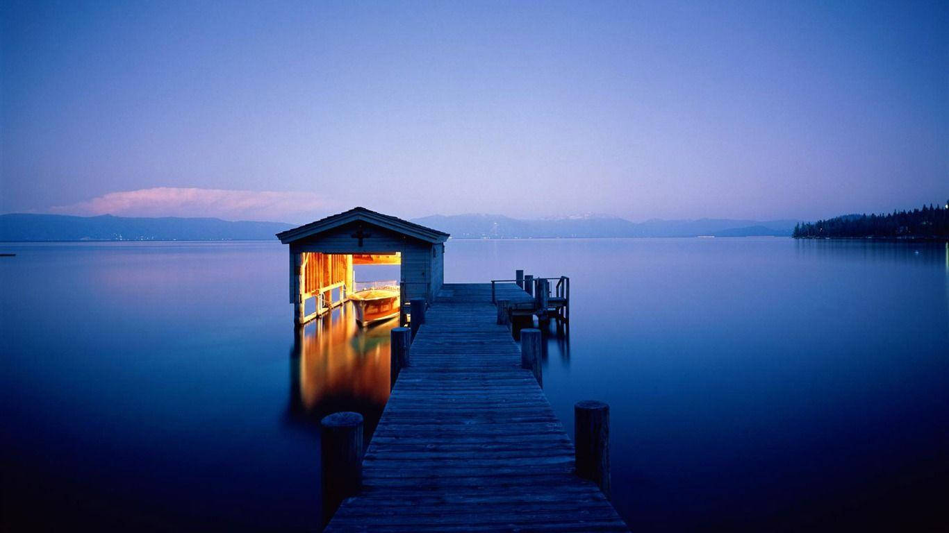 Beautiful Nature Photography 桌布 壁紙 — 一條木製碼頭延伸至暮色中平靜的藍色湖泊，溫暖照亮的船庫倒映在水面上。