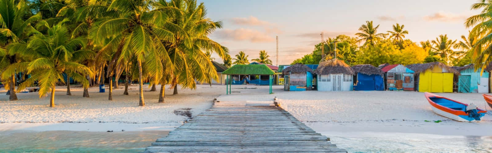 Caribbean Island 桌布 壁紙 — 一條木製碼頭延伸至色彩繽紛的沙灘小屋，在金色日落光芒下，搖曳的棕櫚樹構成畫框。