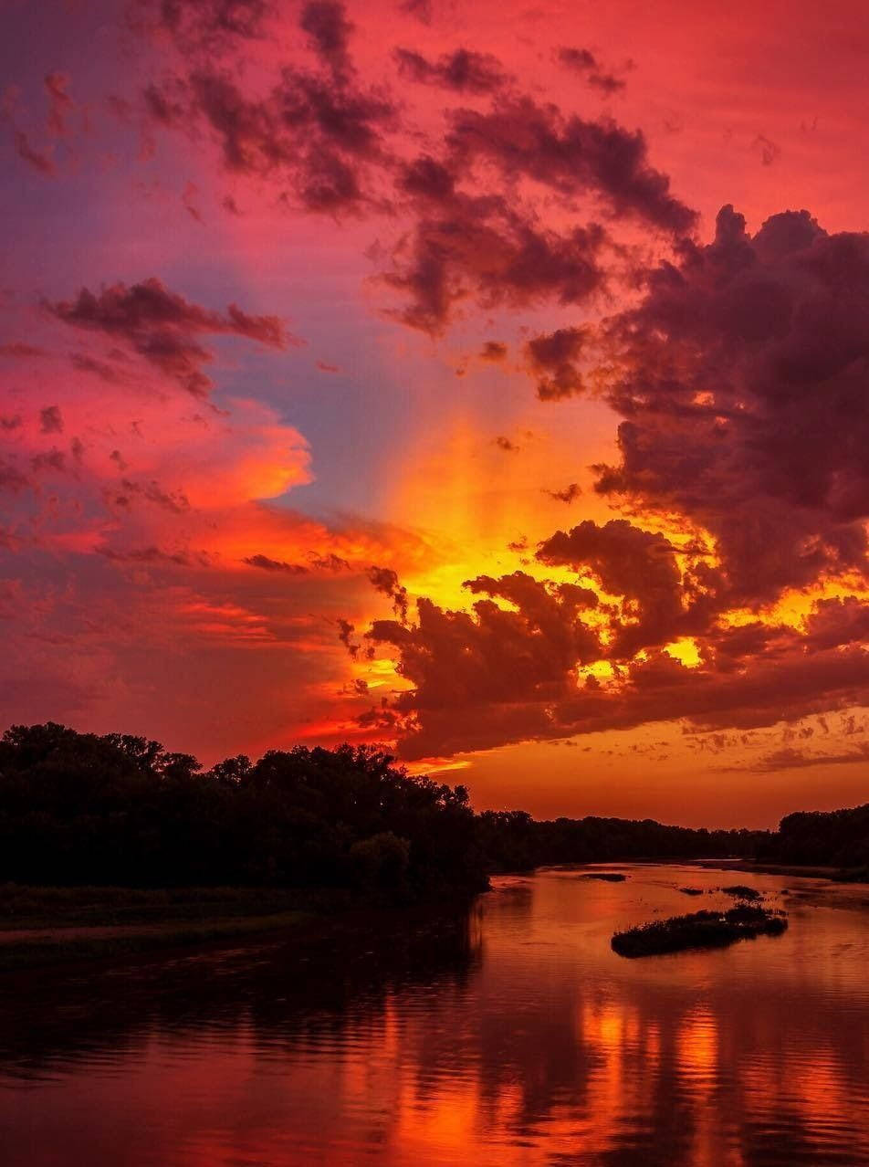 Aesthetic Sunset 桌布 壁紙 — 一條河流映照著火紅的日落，雲朵在黑暗的樹林上方散發橙色和粉紅色的光芒。