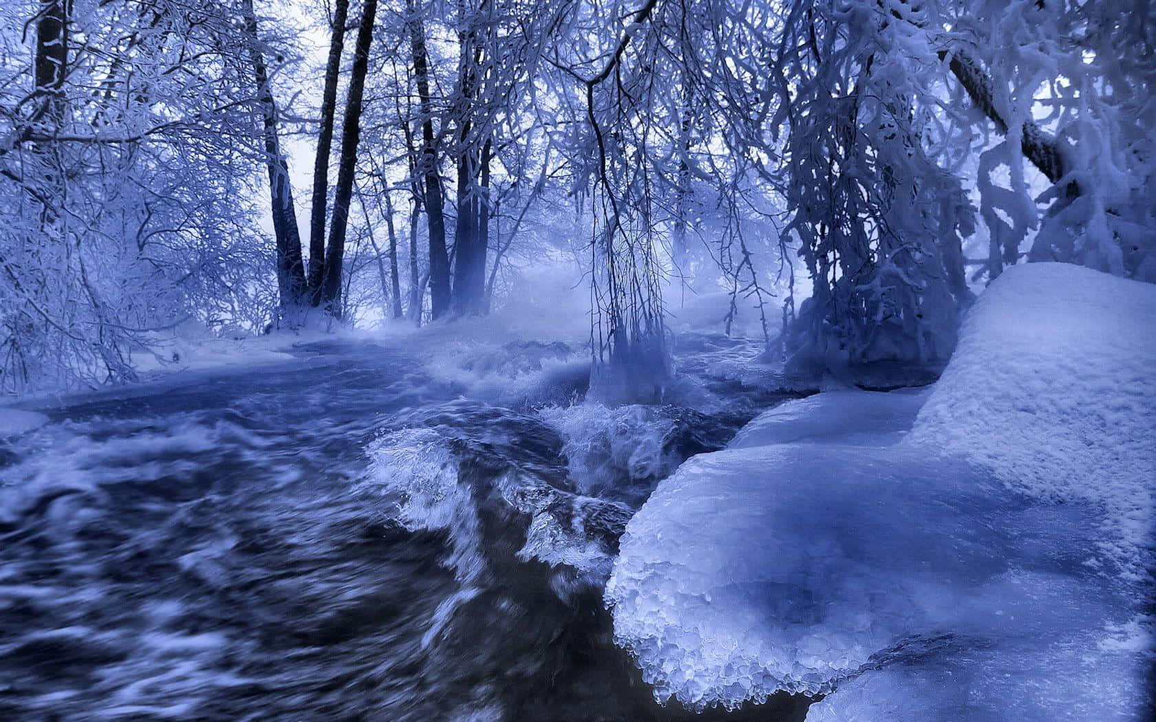 winter solstice 桌布 壁紙 — 一條湍急的冰溪在朦朧的藍色冬日天空下，穿過積雪的森林。