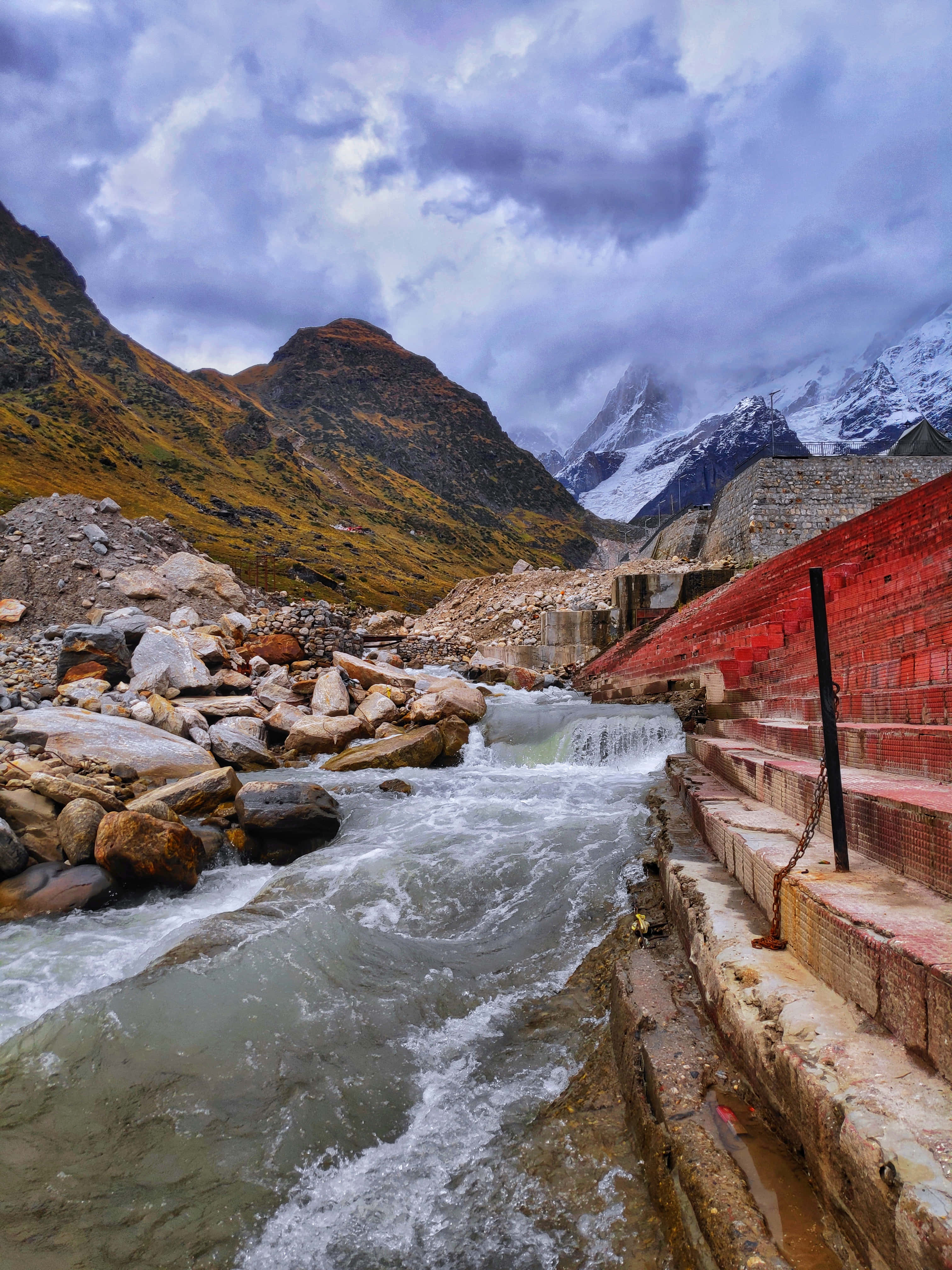 Kedarnath 桌布 壁紙 — 一條湍急的山間溪流流經一座紅磚建築，背景是戲劇性的多雲天空和遠處的雪山頂峰。