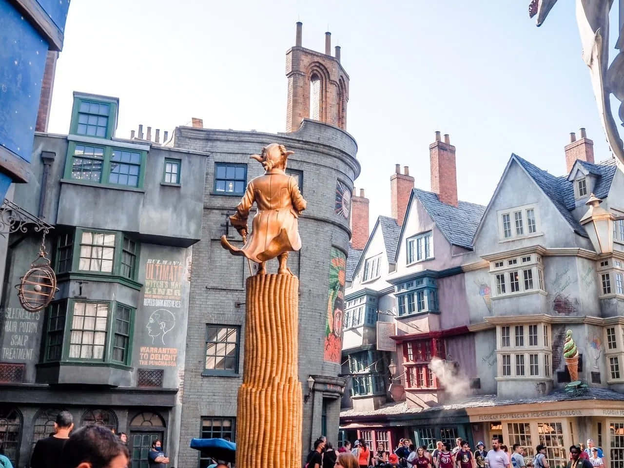 Wizarding World 桌布 壁紙 — 一條熙熙攘攘的巫師街道，高柱上有一座金色女巫雕像，周圍是色彩繽紛的歐式建築。