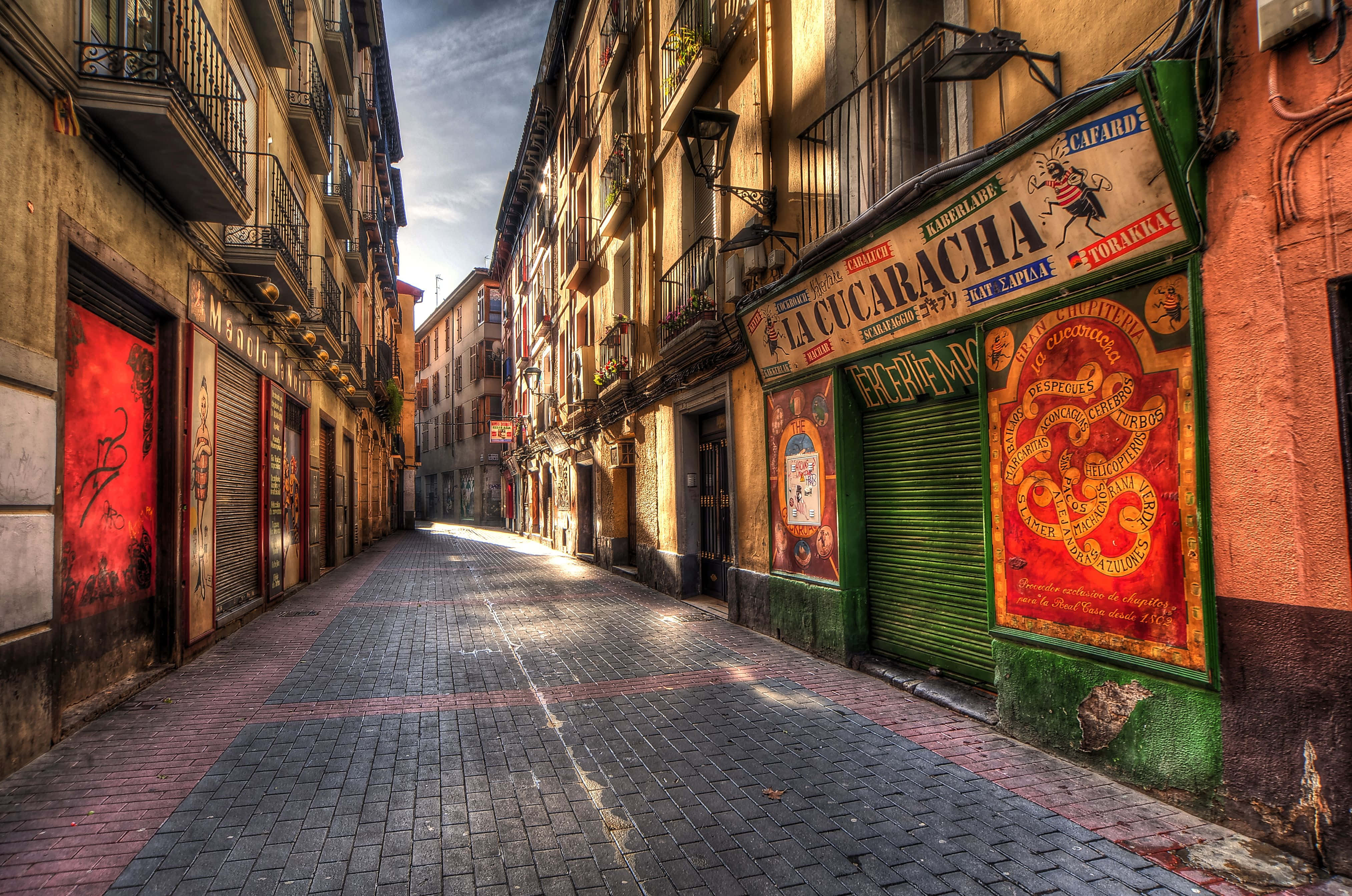 Alleyway 桌布 壁紙 — 一條狹窄、陽光普照的歐洲小巷，有風化的建築物、鮮豔的紅色塗鴉和色彩繽紛的復古煙草店招牌。