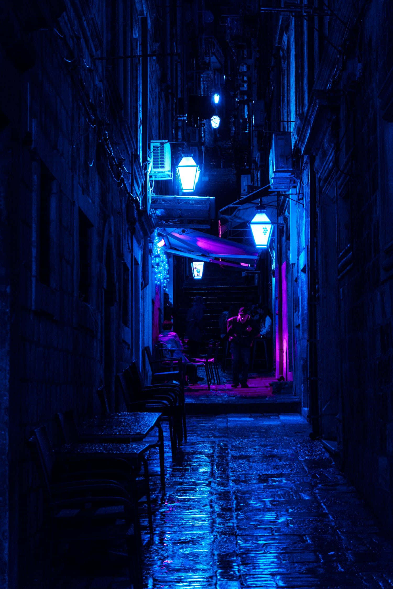 Alleyway 桌布 壁紙 — 一條狹窄潮濕的小巷在夜晚被電藍色霓虹燈照亮，反射在鵝卵石上，戶外餐桌旁有剪影人物。