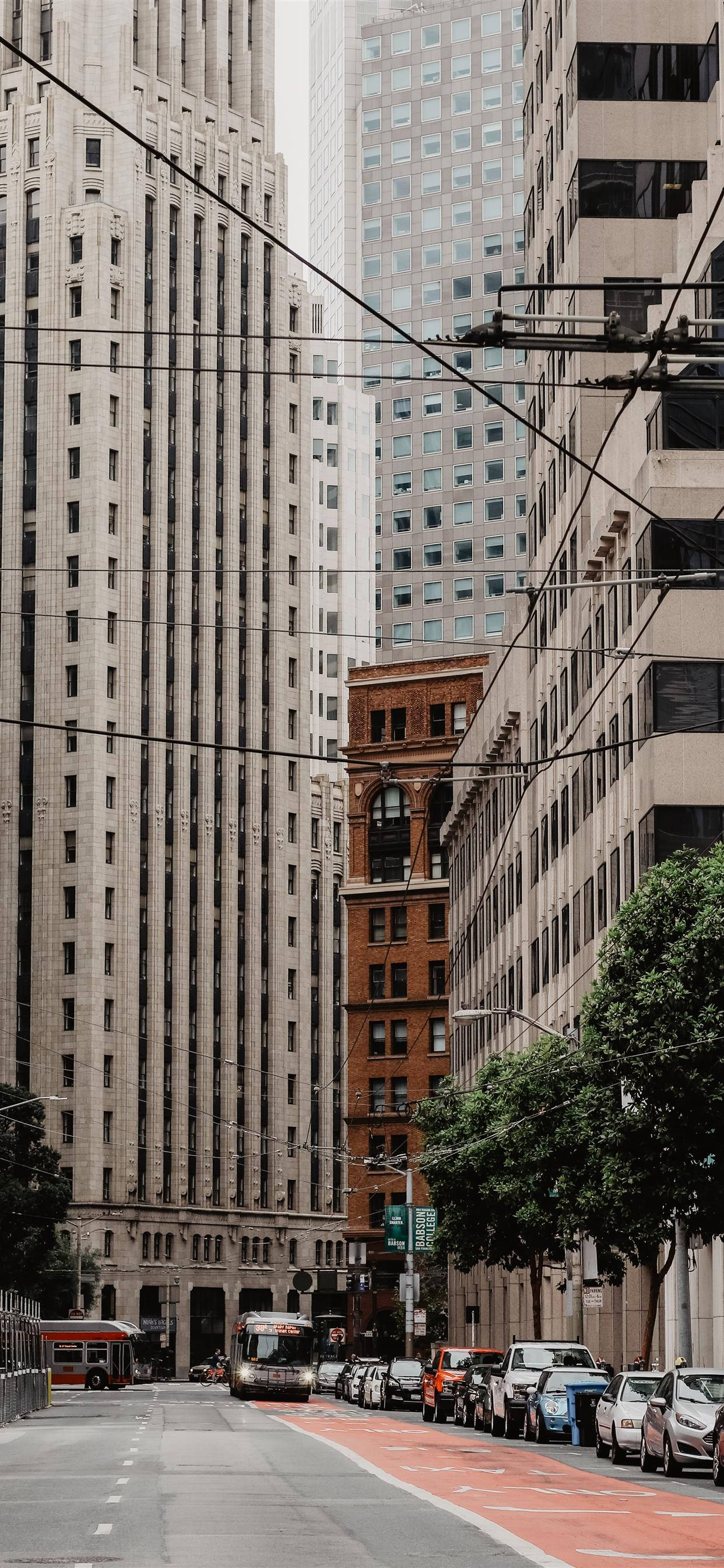 San Francisco Phone 桌布 壁紙 — 一條狹窄的城市街道，兩旁是高聳的摩天大樓和歷史悠久的磚砌建築，一輛纜車從架空電線下經過。