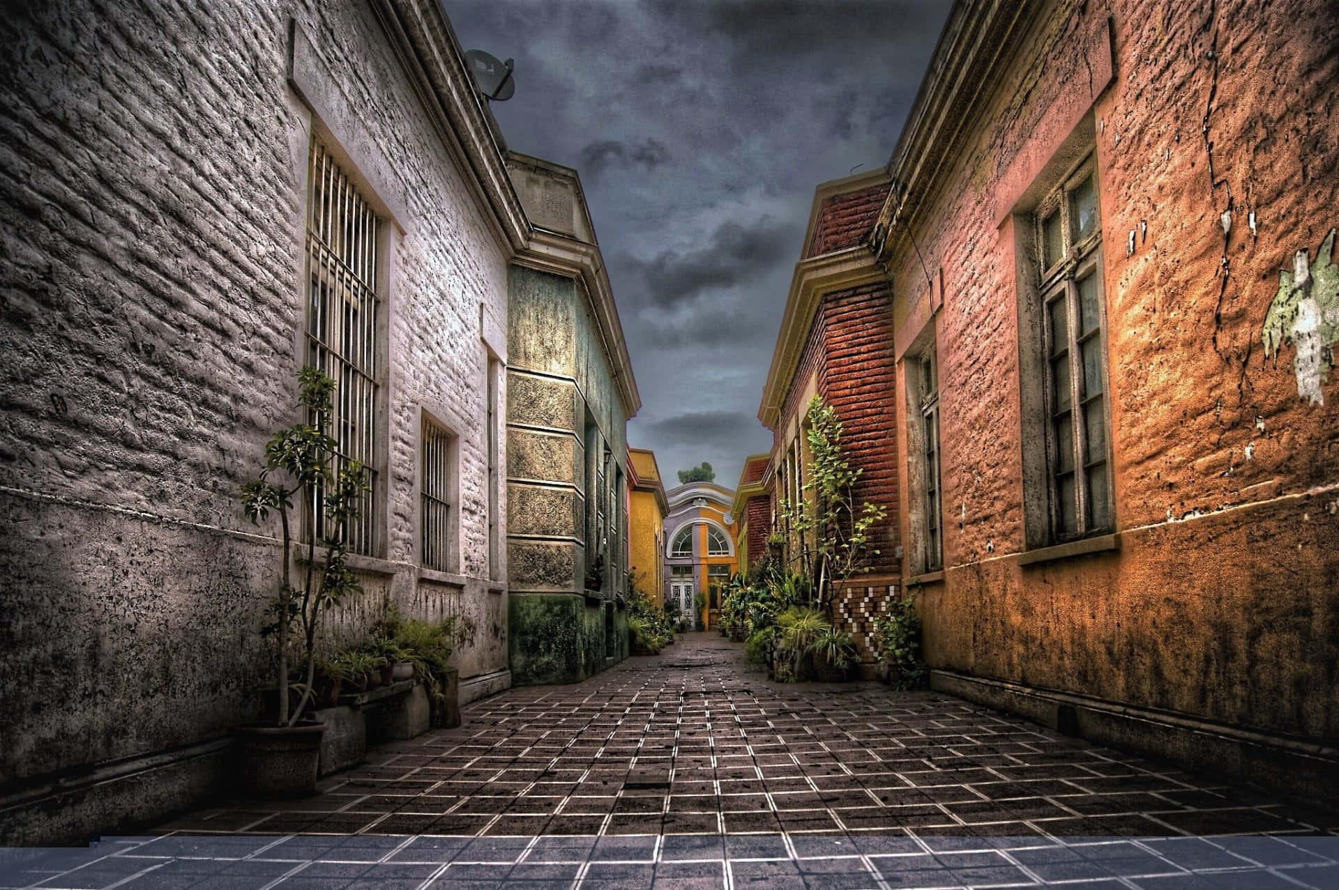 Alleyway 桌布 壁紙 — 一條狹窄的小巷，兩旁是風化的磚砌建築，在暴風雨的天空下，盆栽植物排列在鵝卵石小徑上。