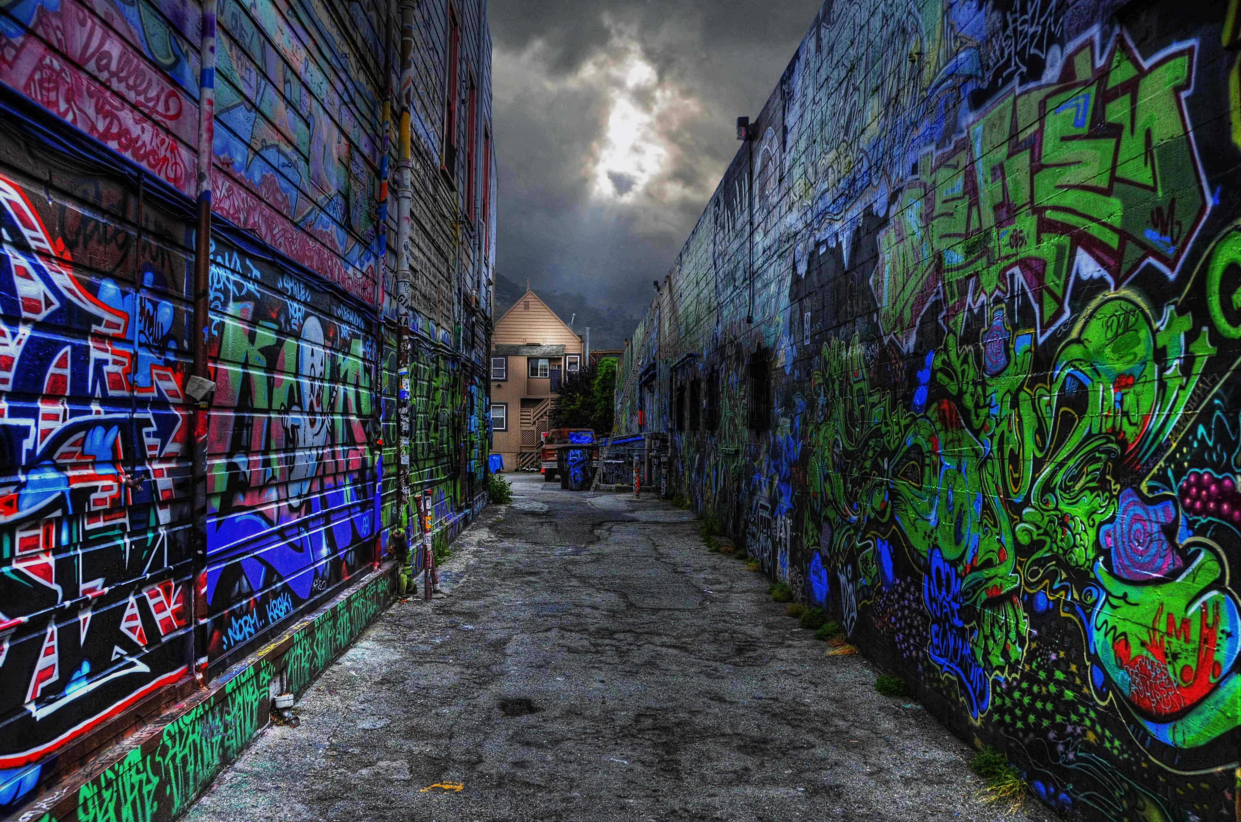Alleyway 桌布 壁紙 — 一條狹窄的小巷，兩旁牆壁在戲劇性的多雲天空下佈滿鮮豔的塗鴉。