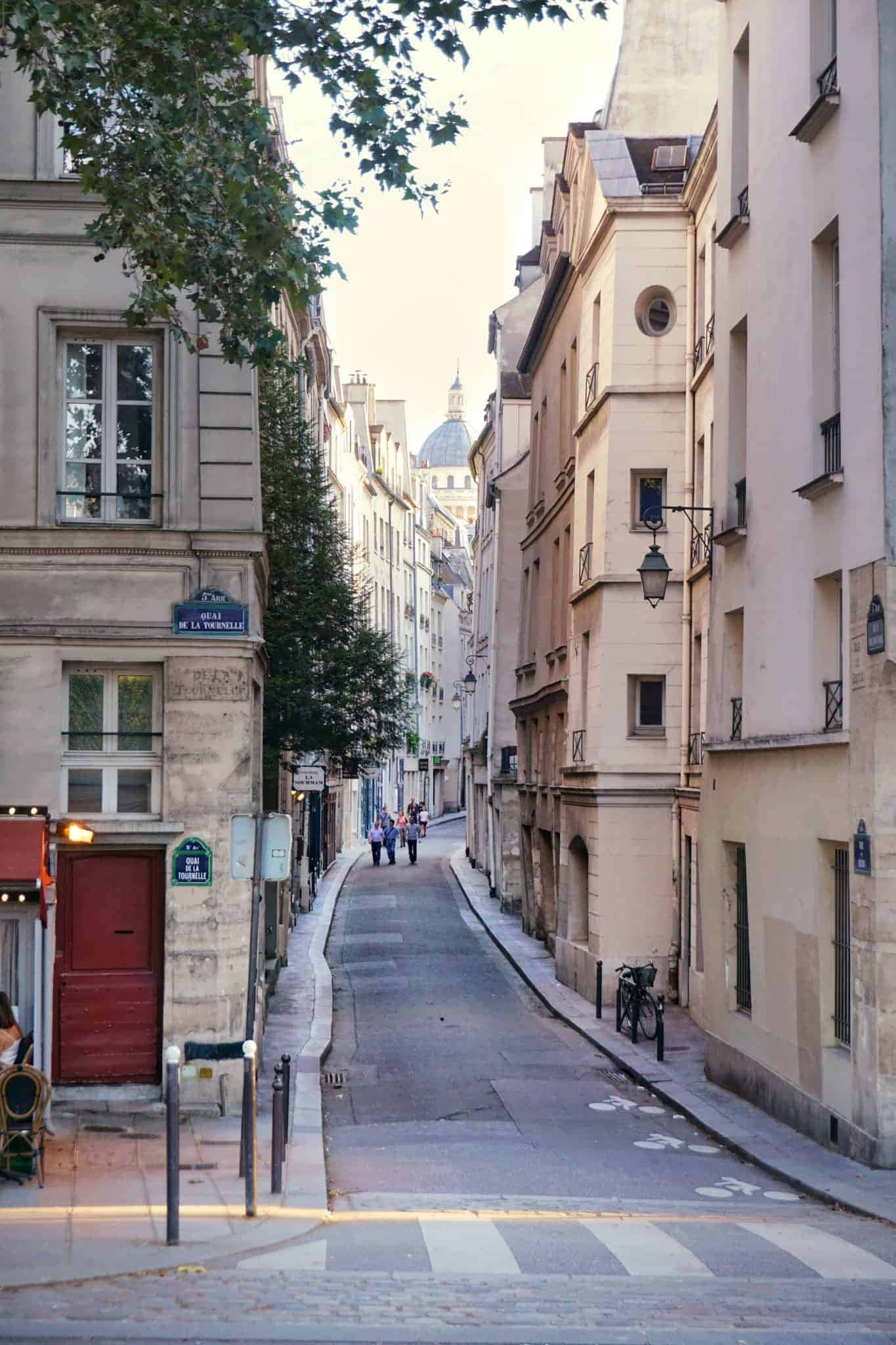 Street 桌布 壁紙 — 一條狹窄的巴黎街道，兩旁是歷史建築和樹木，在柔和日光下通往遠處的圓頂大教堂。