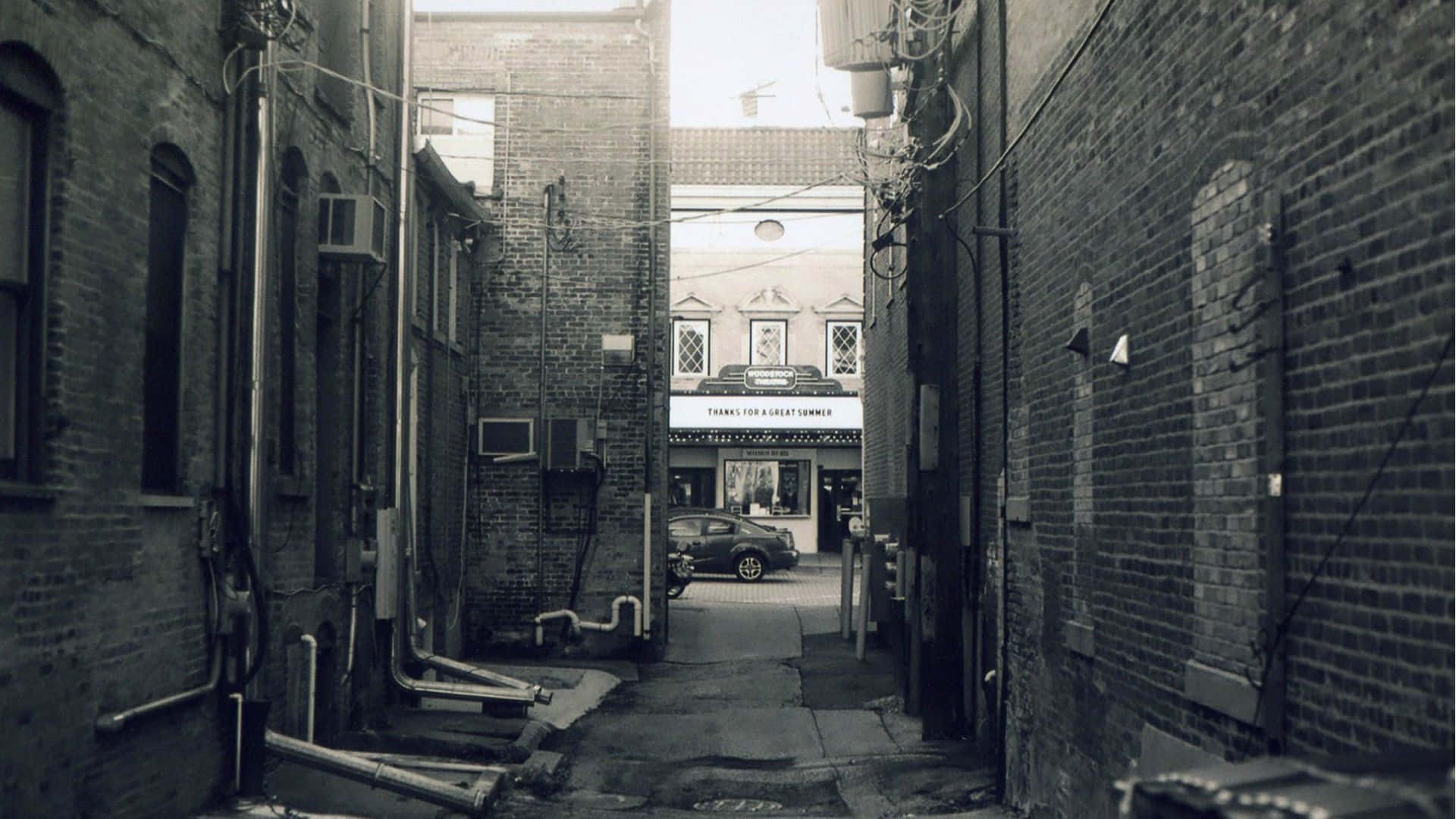Alleyway 桌布 壁紙 — 一條狹窄的磚砌小巷通向一個復古店面，店前停著一輛車，天空明亮。